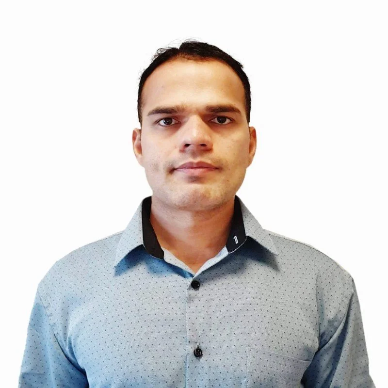 A man with short dark hair, wearing a gray collared shirt with black accents, standing against a white background.