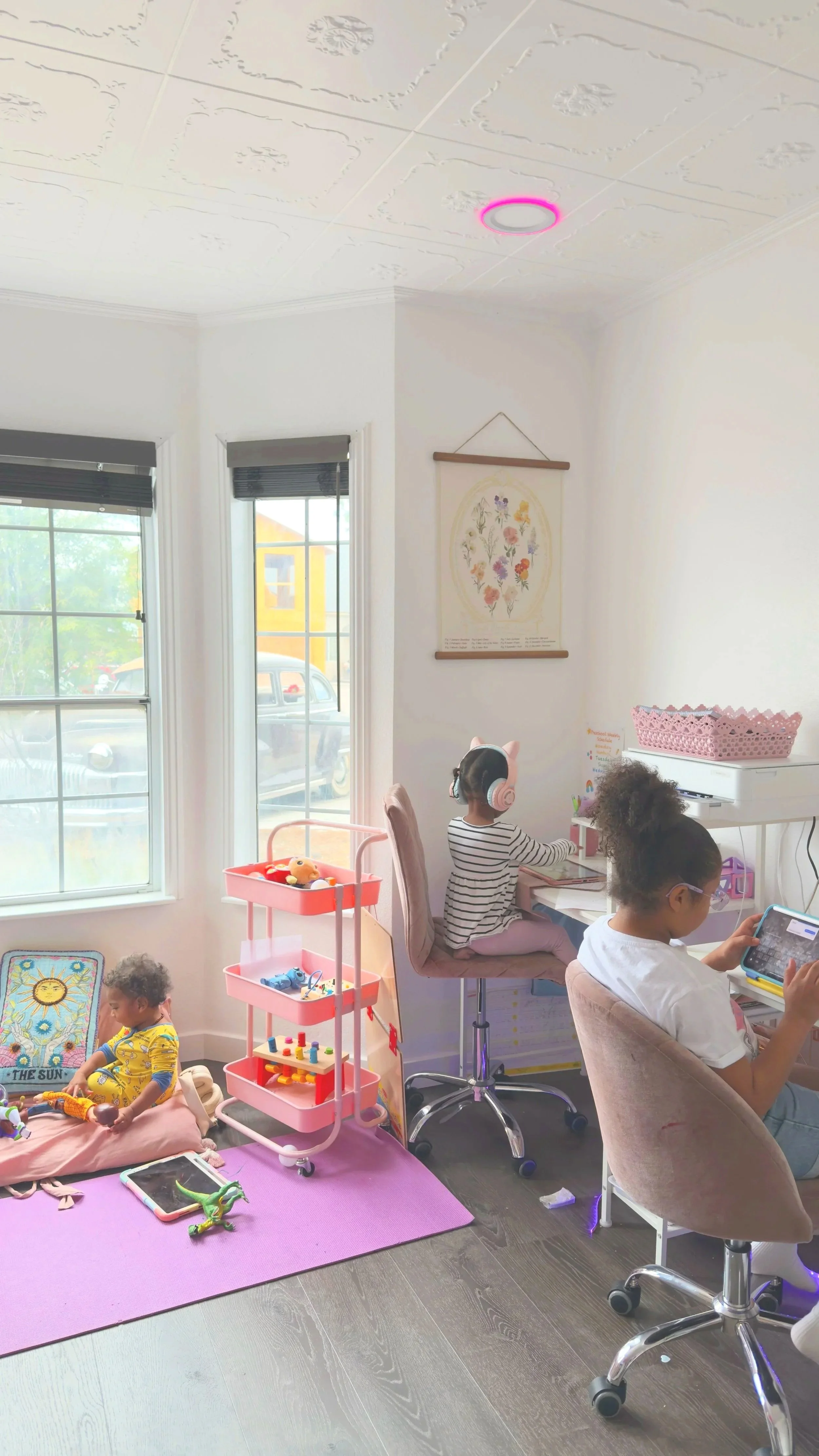 A room with three children, two adults, and various toys and furniture. One child is sitting on a pink mat playing with toys. Another child is at a desk with headphones, and the third is seated in a chair using a tablet. There are large windows, a pink toy cart, a framed picture of the sun, and a white piano with a pink basket on top.
