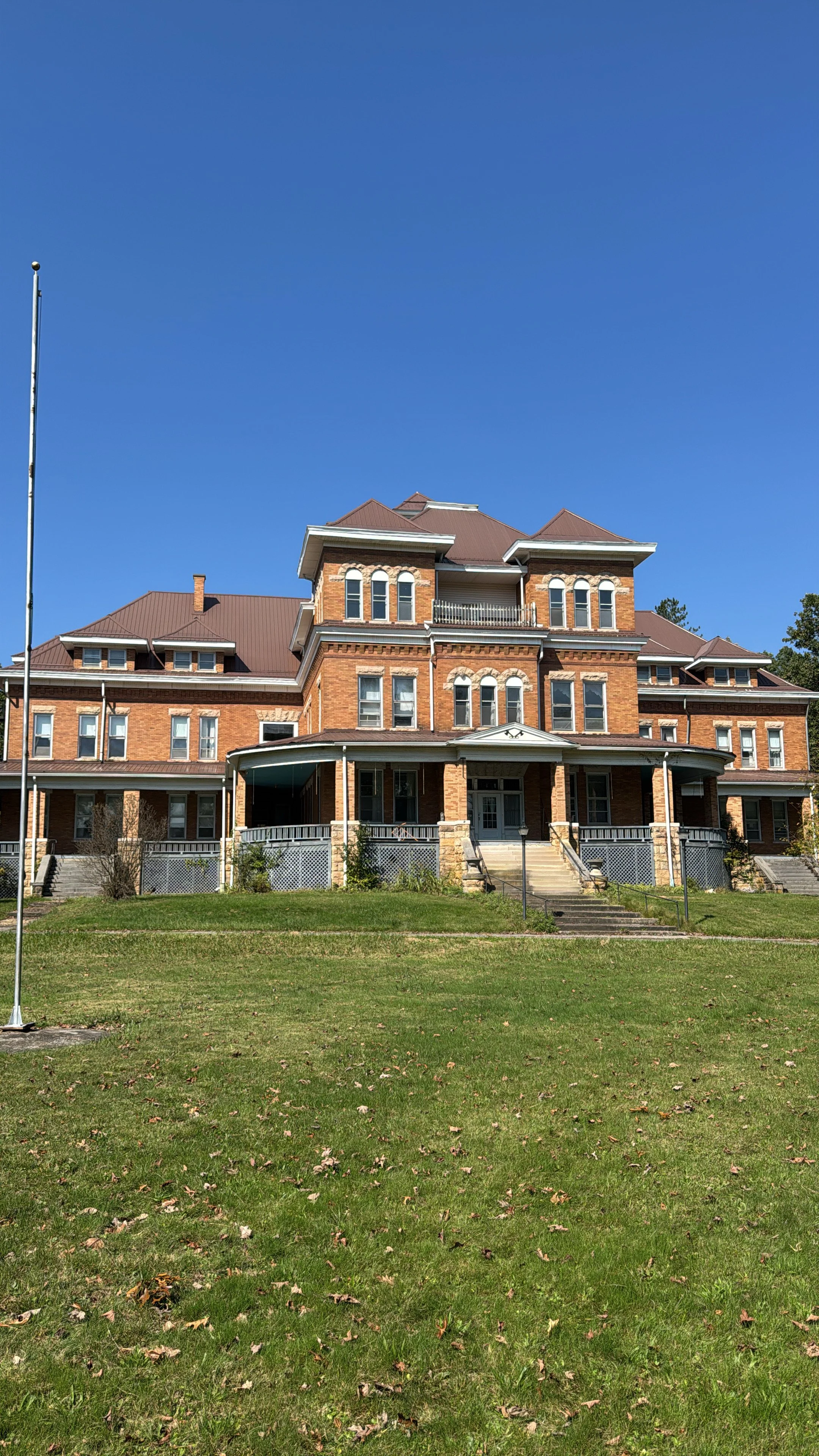 Large historic brick building with multiple stories, a wrap-around porch, and a steep roof, set against a bright blue sky and green lawn in front.