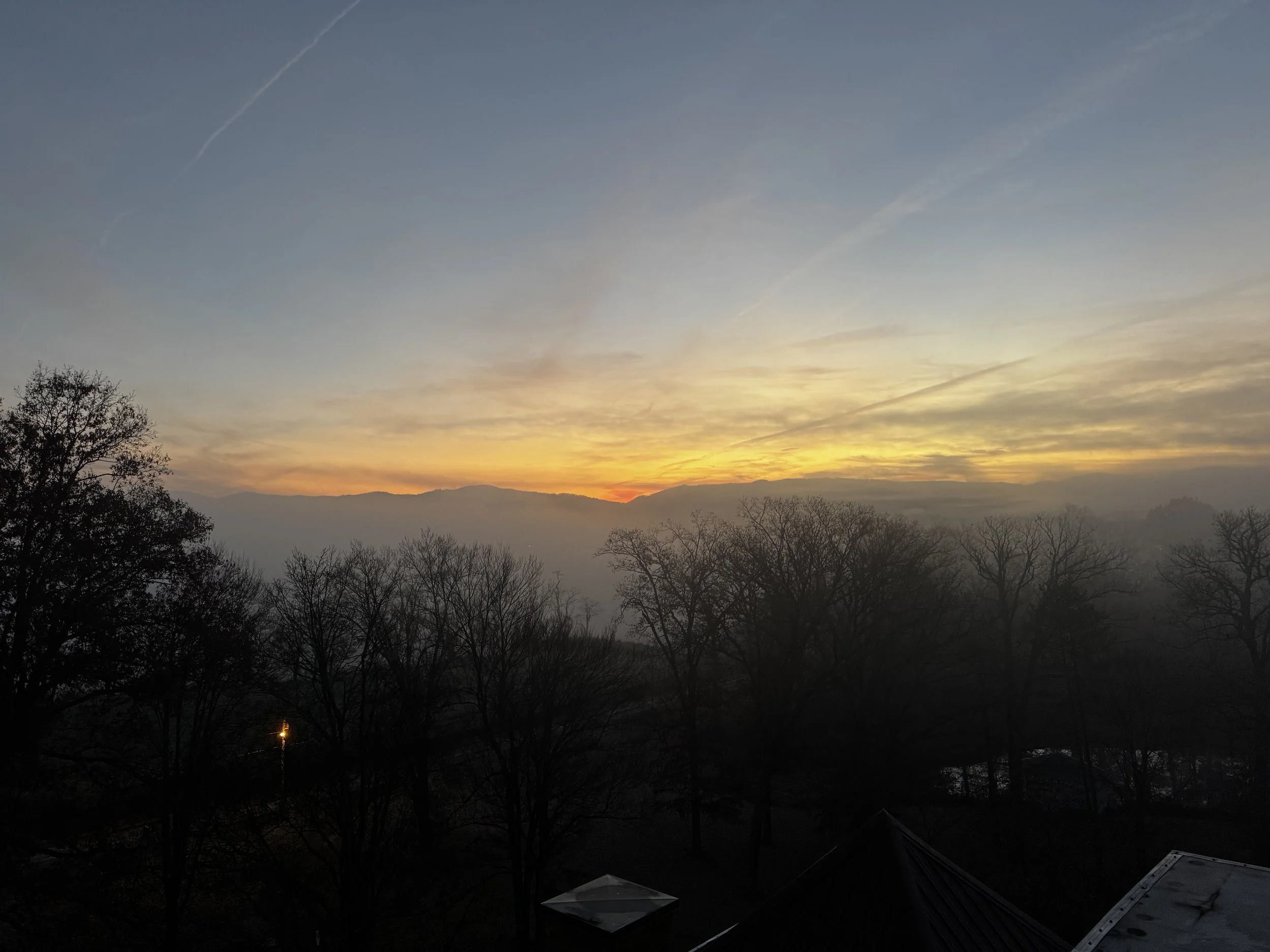 Sunrise over a mountain range with a sky filled with clouds and contrails, with silhouetted trees and rooftops in the foreground.