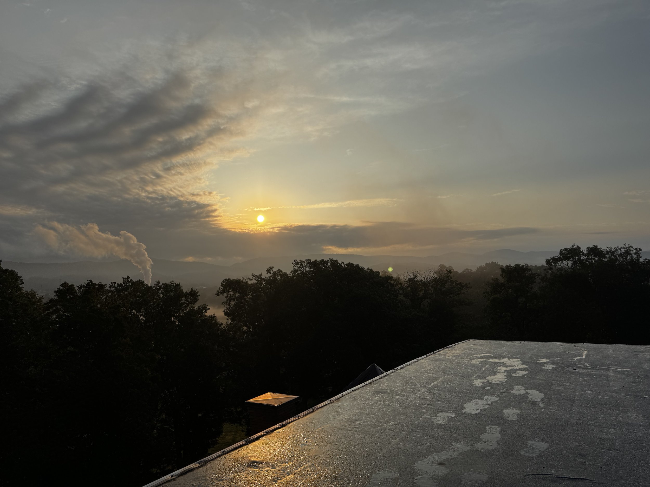 Golden sunset over layered mountains and silhouetted trees, viewed from a rain-speckled rooftop beneath dramatic clouds