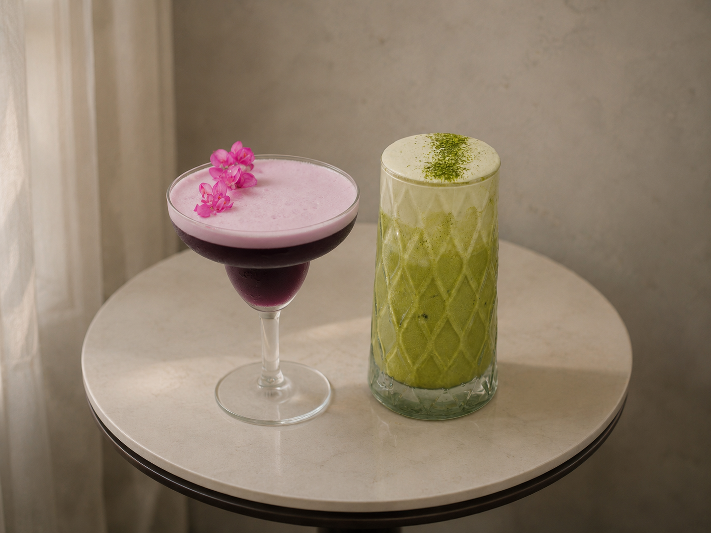 Two cocktails on a round table, one with pink liquid and floral garnish in a coupe glass, the other with green liquid and powdered garnish in a tall glass.