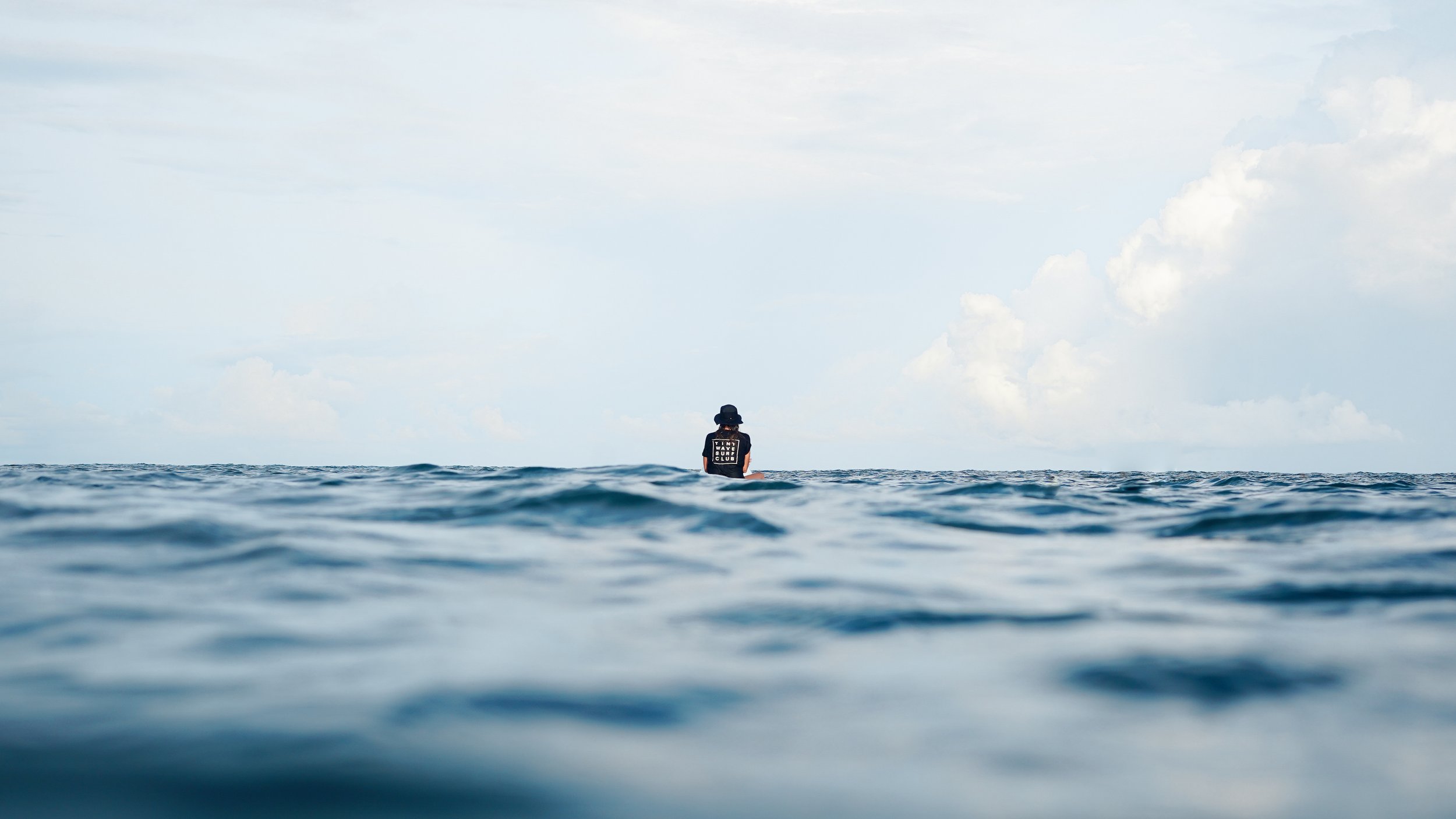 Person wearing the classic logo tee sitting in the ocean, with a cloudy sky in the background.