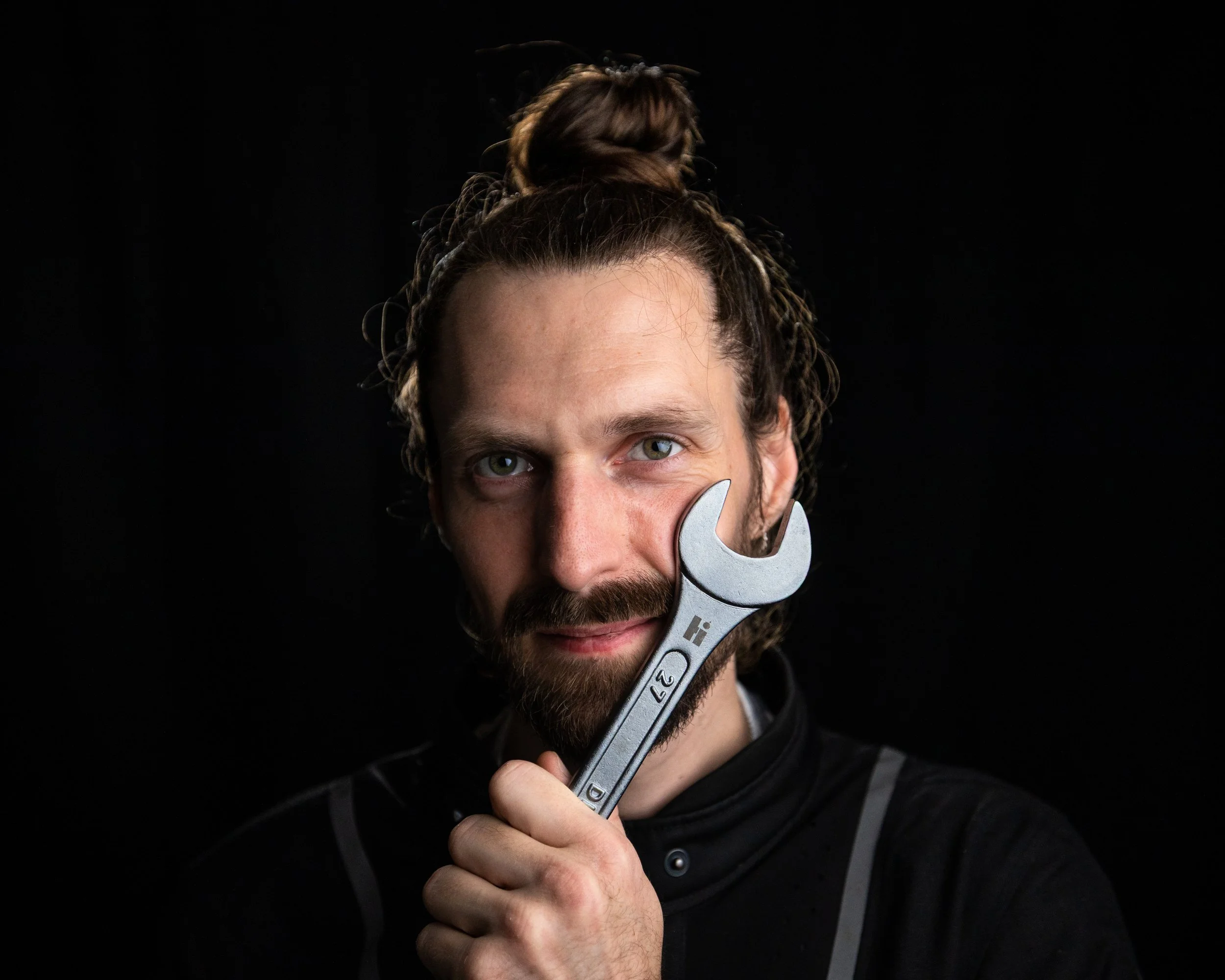 A man with long hair and a beard holding a large wrench up to his face, partially covering his cheek, against a black background.