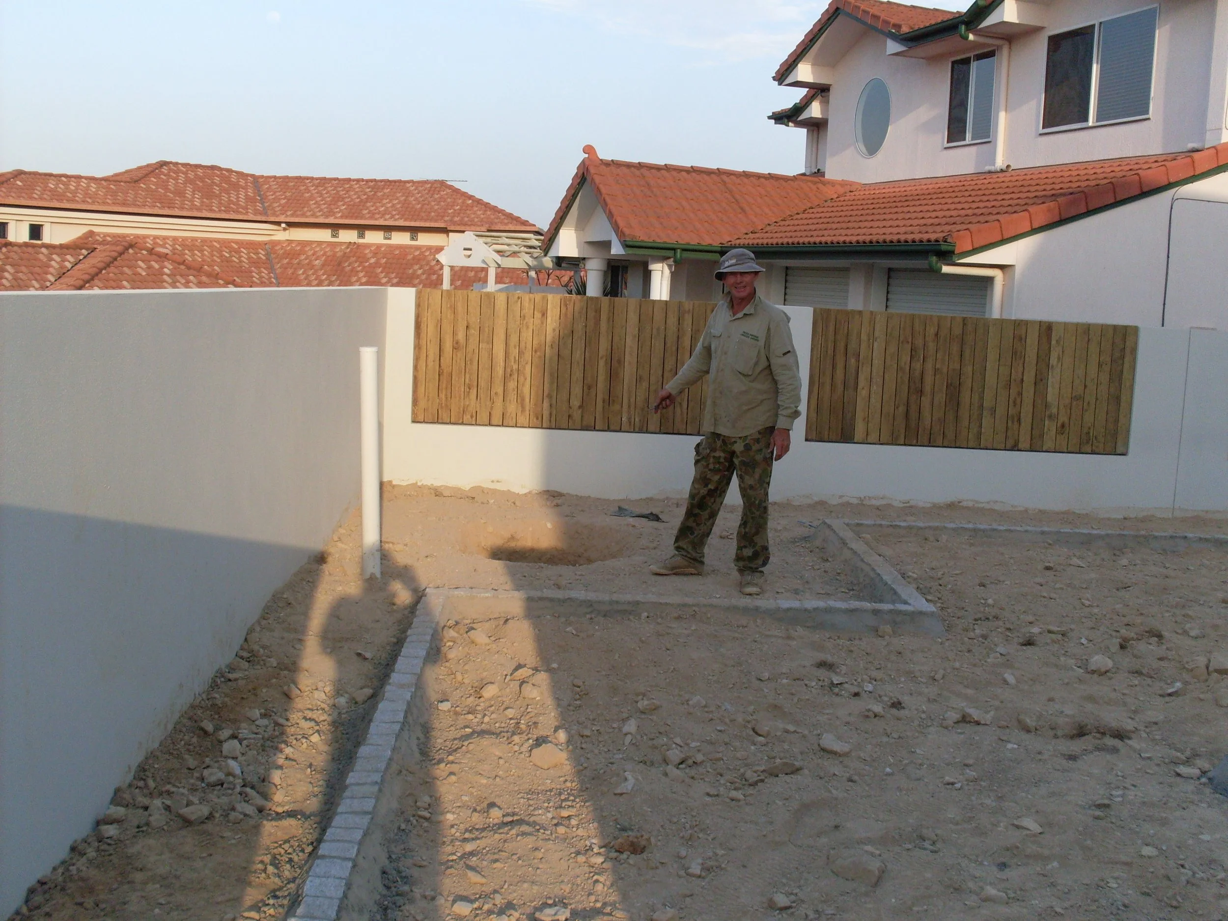 Man standing on a construction site with partially built walls and tiled-roof houses in the background