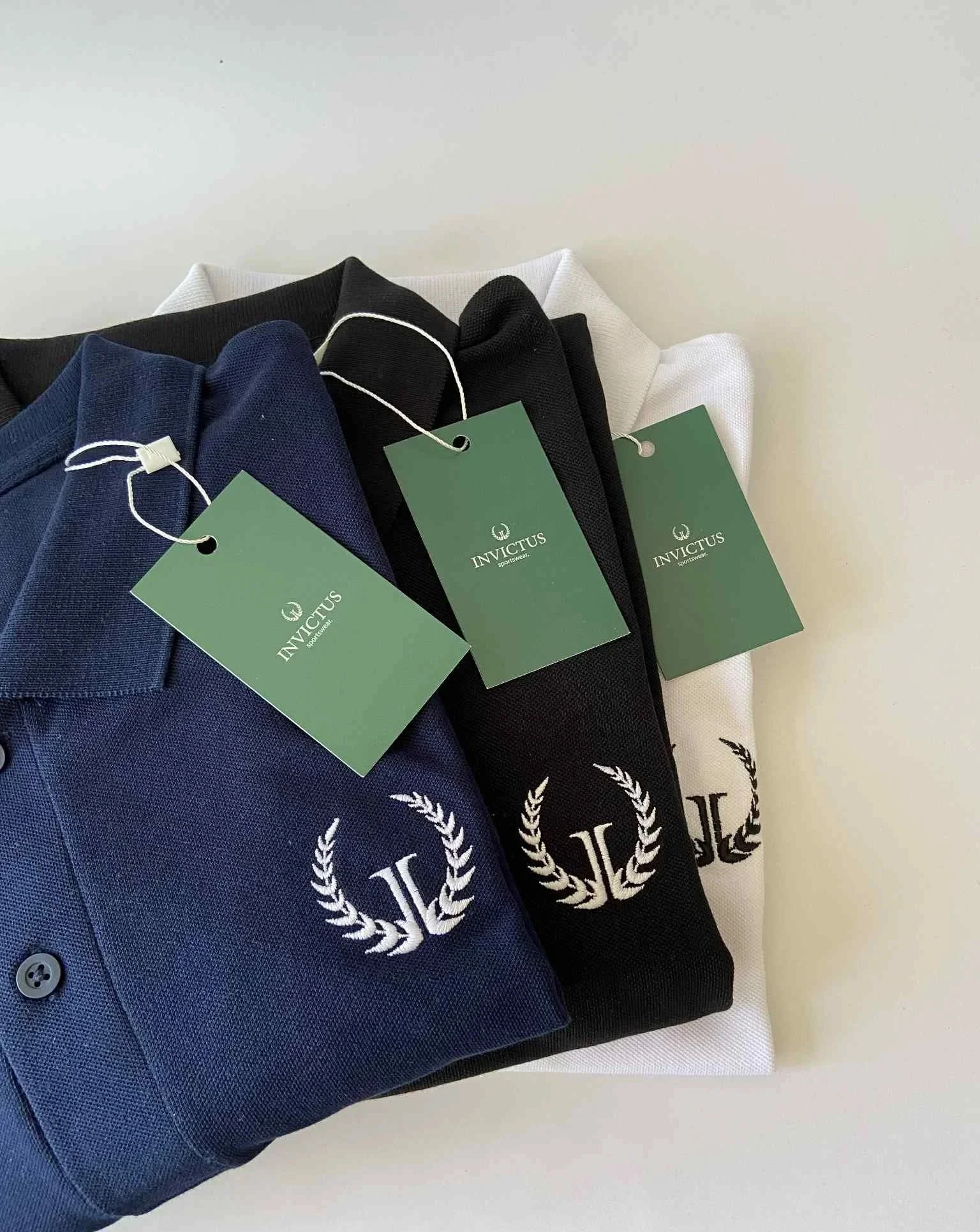 Folded polo shirts in navy, black, and white with embroidered logo and tags labeled 'Invictus' on a white surface.