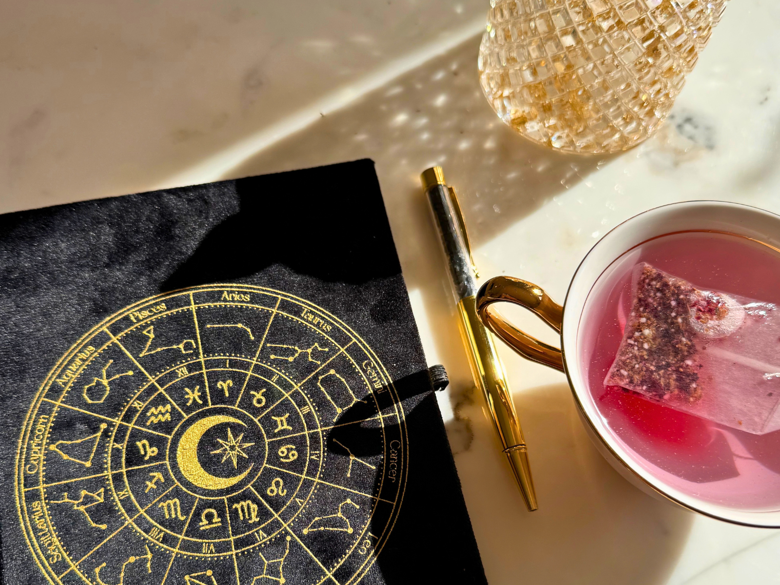 Black velvet astrology journal with a gold pen and a cup of fruit tea in dappled sunlight