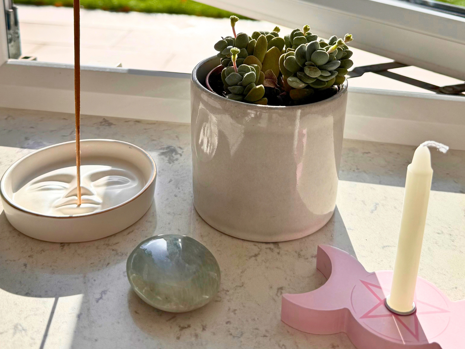 Lit incense in a moon-face holder on a sunny windowsill beside a spell candle