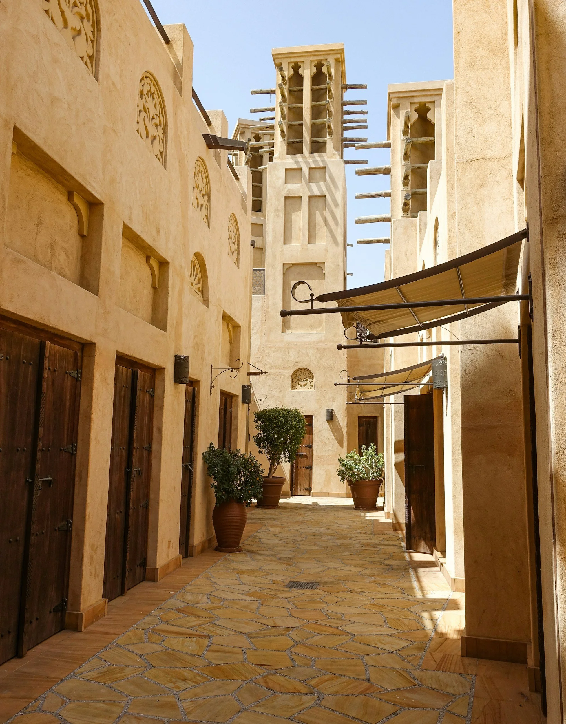 Traditional Middle Eastern architecture with wind towers, narrow alleyways, wooden doors, potted plants, and stone pathway under clear sky. Dubai, United Arab Emirates, UAE