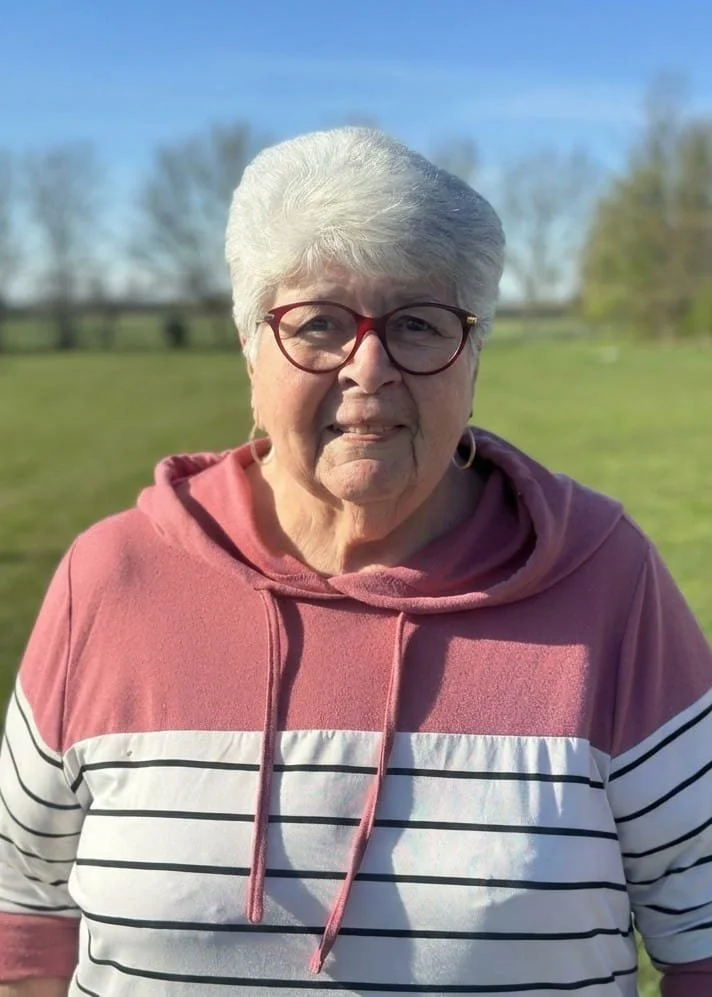Smiling woman with short curly hair and glasses, wearing a black top with a necklace, standing outdoors among greenery and purple flowers.