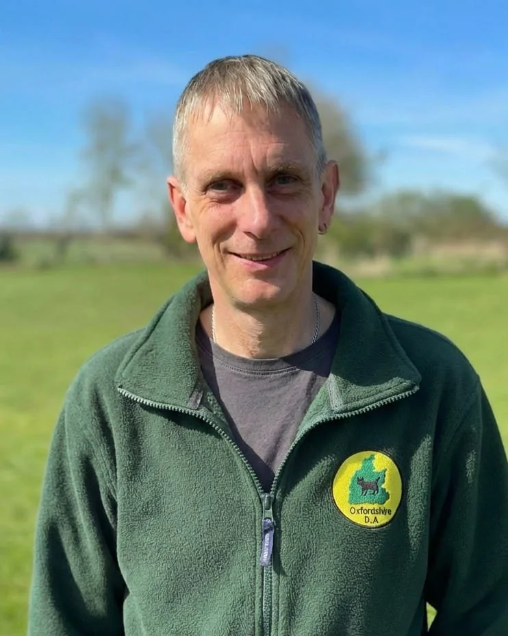A man with light hair and a friendly smile, wearing a blue shirt, outdoors.