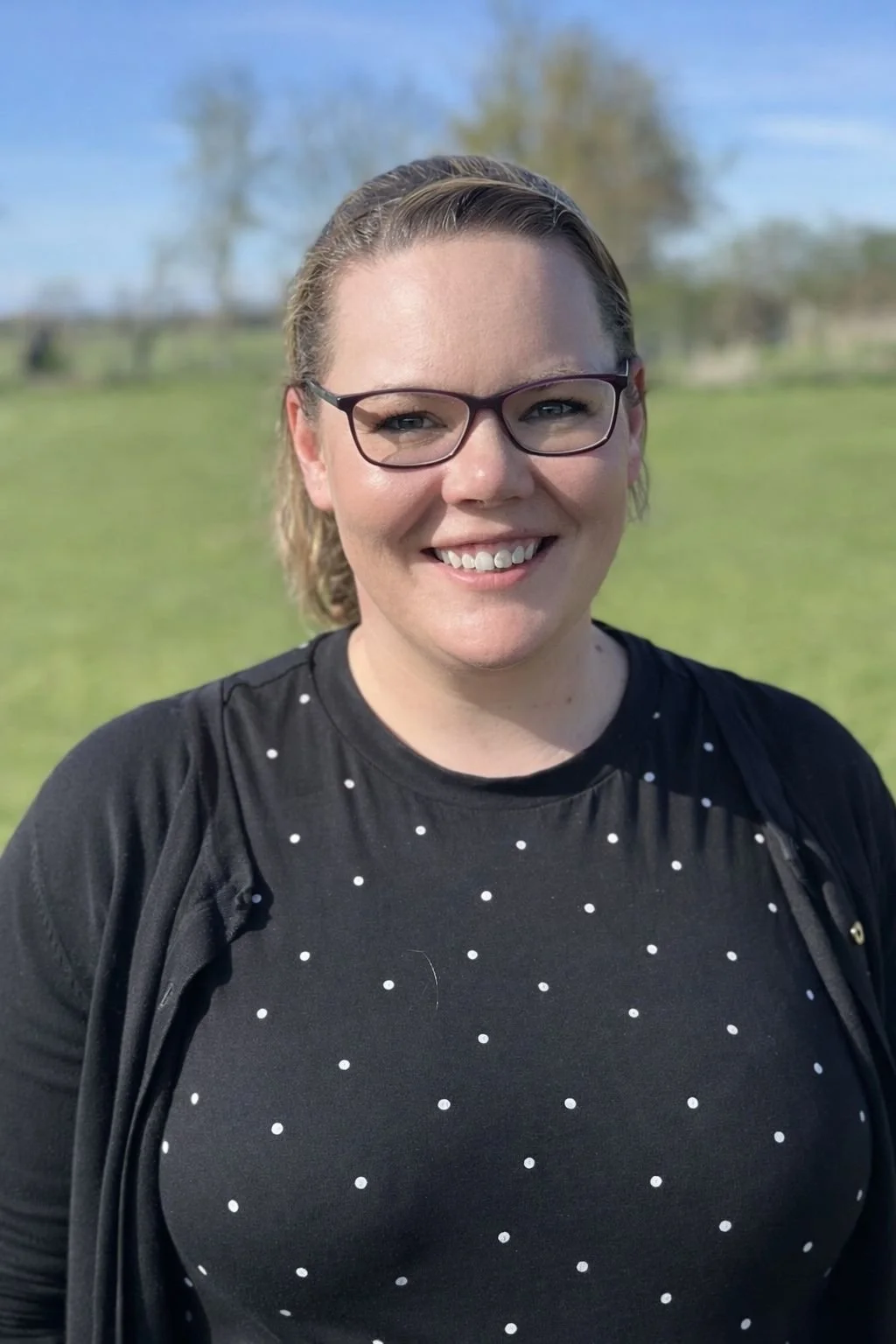 A woman with light brown hair, wearing glasses and a blue sleeveless top, smiling at the camera in an indoor setting.