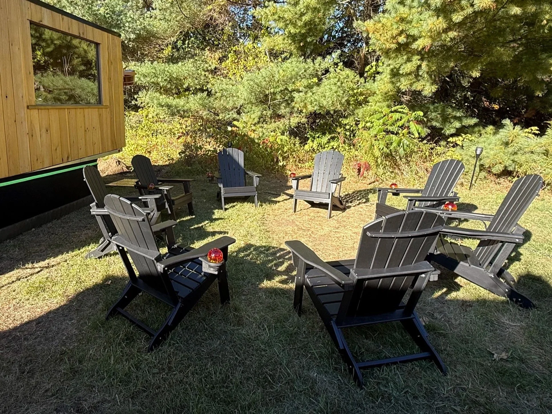 A backyard with eight black Adirondack chairs arranged in a circle on a grassy patch, surrounded by trees and bushes, with one small red lantern or decorative object on each chair's armrest.