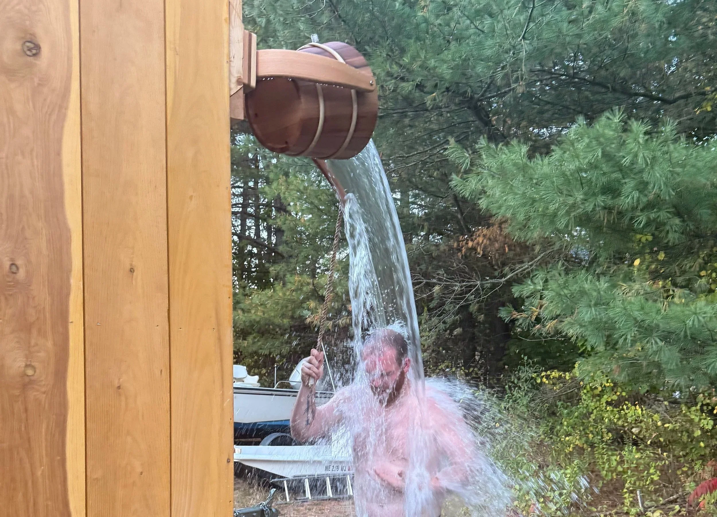 A man is getting splashed with water from a wooden barrel positioned above him. He is holding a rope to tilt the barrel, causing water to pour down. The background shows trees and a boat on a trailer.