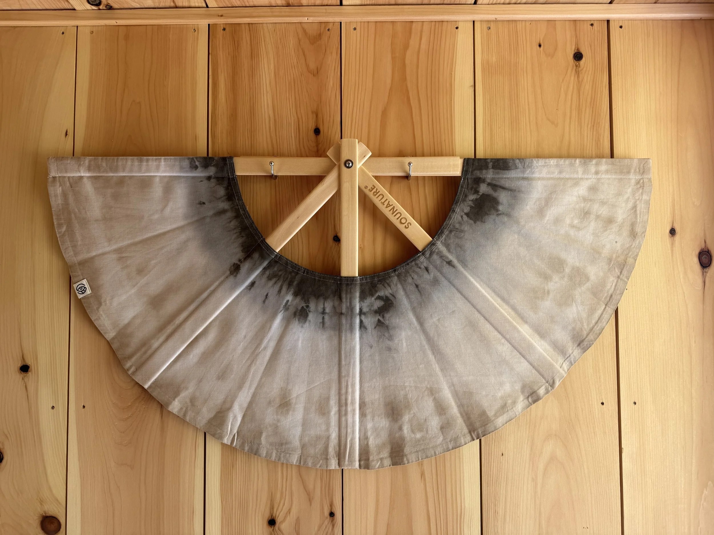 A fan-shaped piece of fabric with black and beige tie-dye pattern, mounted on a wooden wall, with a wooden support labeled 'SOUNAMATURE'.