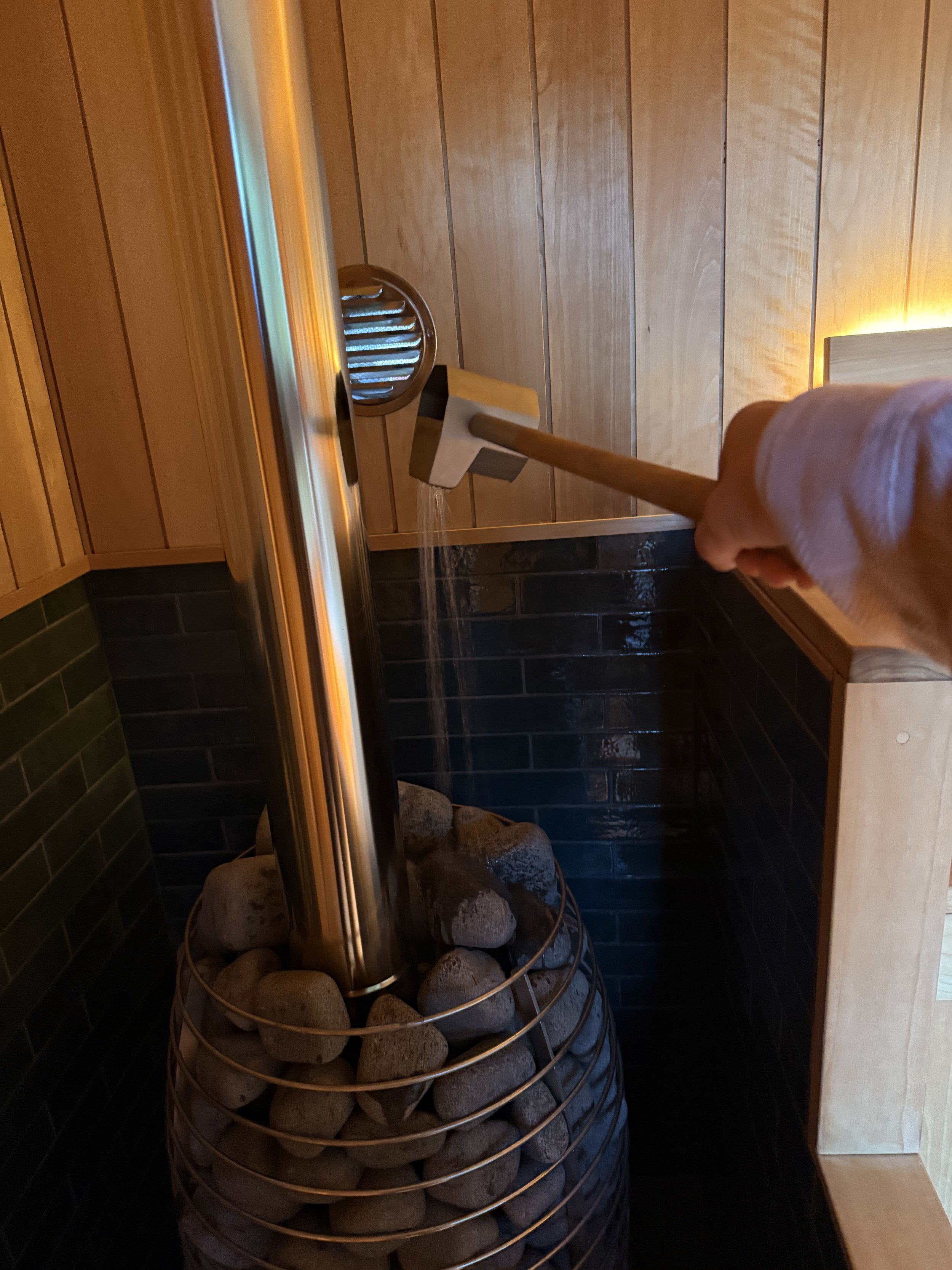 A hand holding a wooden ladle pouring water over hot stones in a sauna heater.