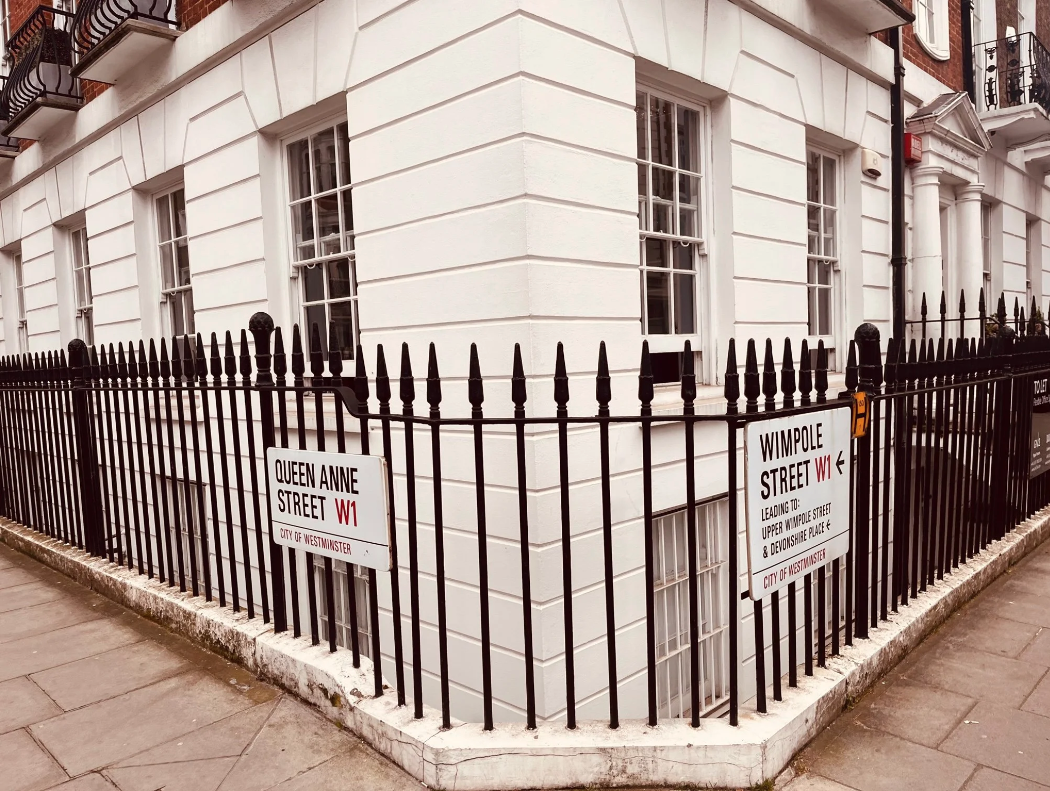 A corner of a building with white painted brick style facade, surrounded by a black metal fence with pointed tops. There are multiple signs on the fence indicating walking directions, including Queen Anne Street W1 and Wimpole Street W1 in Westminster, London. The building has bay windows, decorative columns at the entrance, and fire escape balconies.