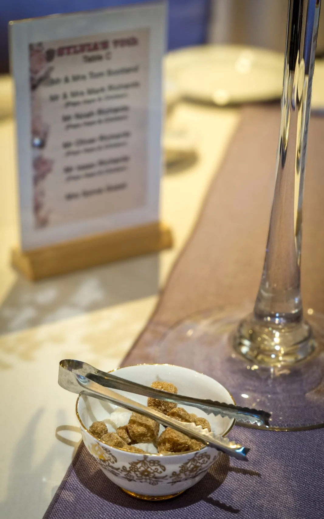 A small white bowl with gold patterns filled with brown sugar cubes, with silver tongs resting on top, on a table with a purple tablecloth. In the background, a menu and a large empty wine glass are visible.