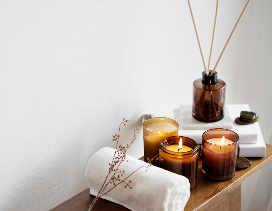 A spa setup with burning candles, aromatic reed diffuser in brown glass, a white rolled towel, and decorative stones on a wooden surface against a white backdrop.