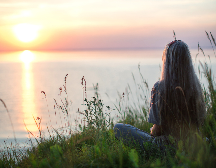 Person sitting on a grassy hill overlooking a sunset by the water.