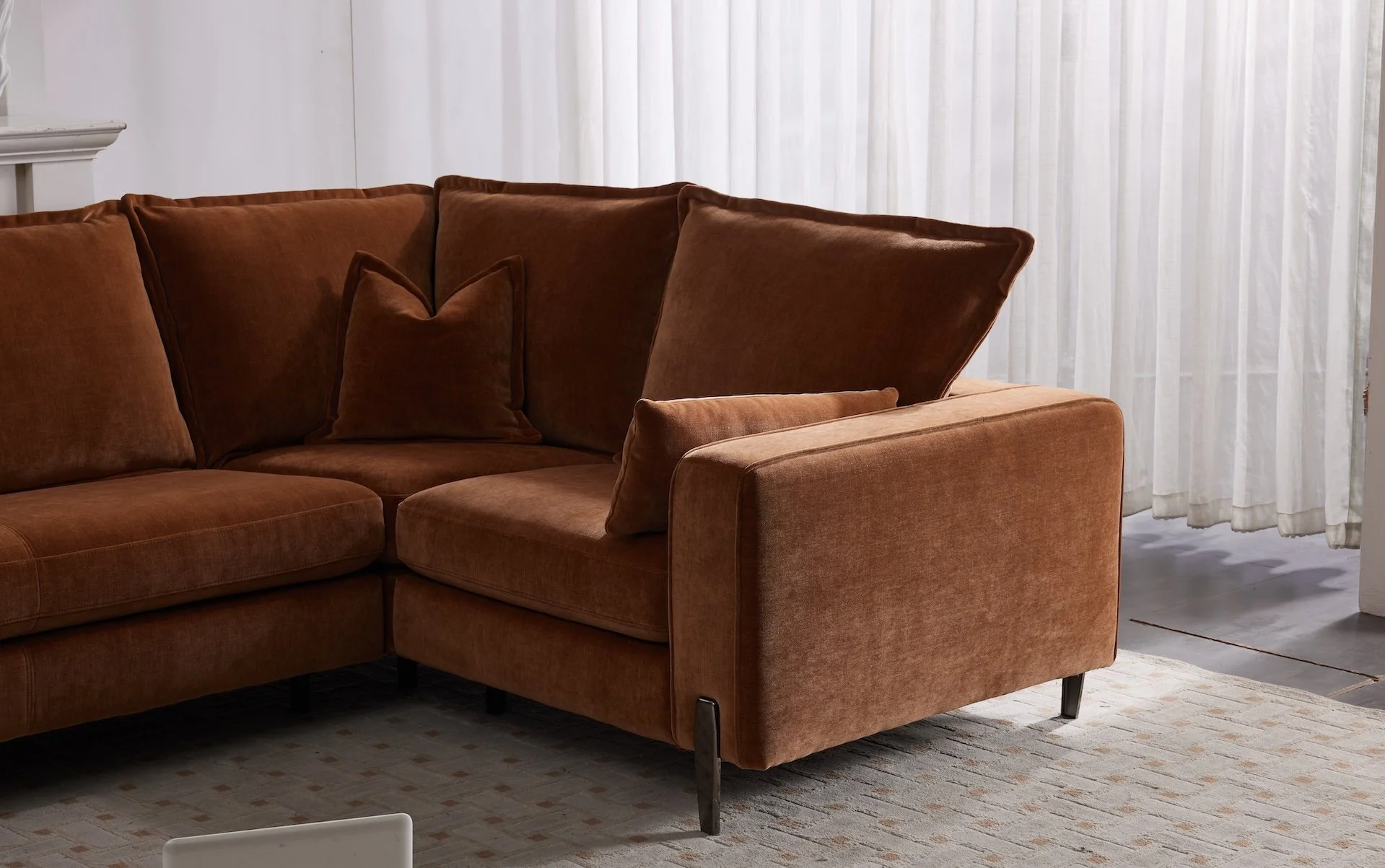 Brown sectional sofa with cushions in a living room with white curtains and a patterned rug.