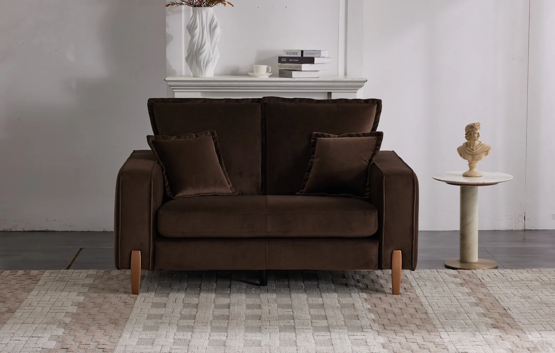 A room with a brown velvet loveseat sofa with two matching pillows, a white mantel shelf with decorative items including a sculpture, books, and a cup, and a small round side table with a bust sculpture on top. The floor is covered with a beige textu