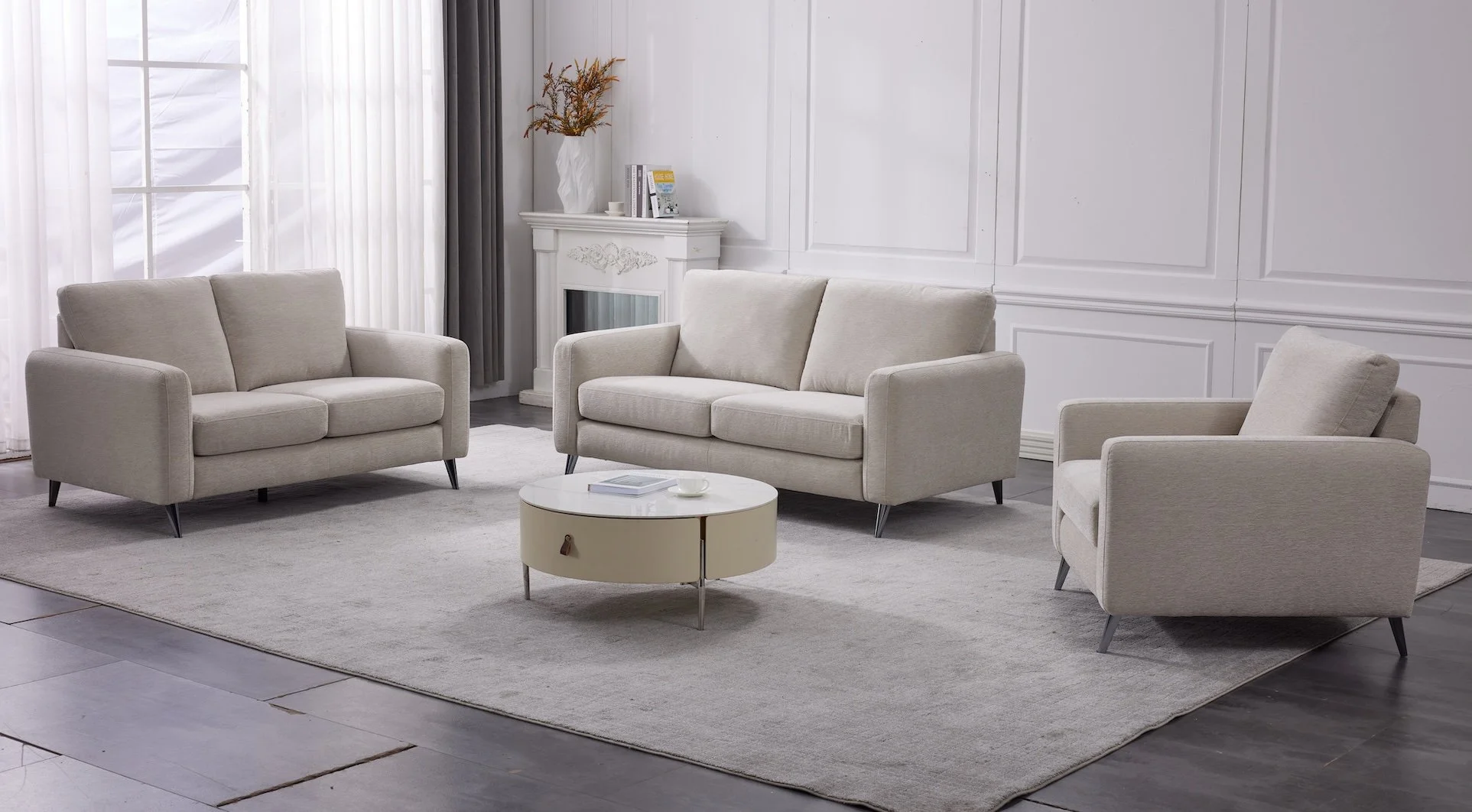 A living room with three beige upholstered sofas, a round white coffee table, a white fireplace mantel with decor, a large window with curtains, and gray-painted walls.