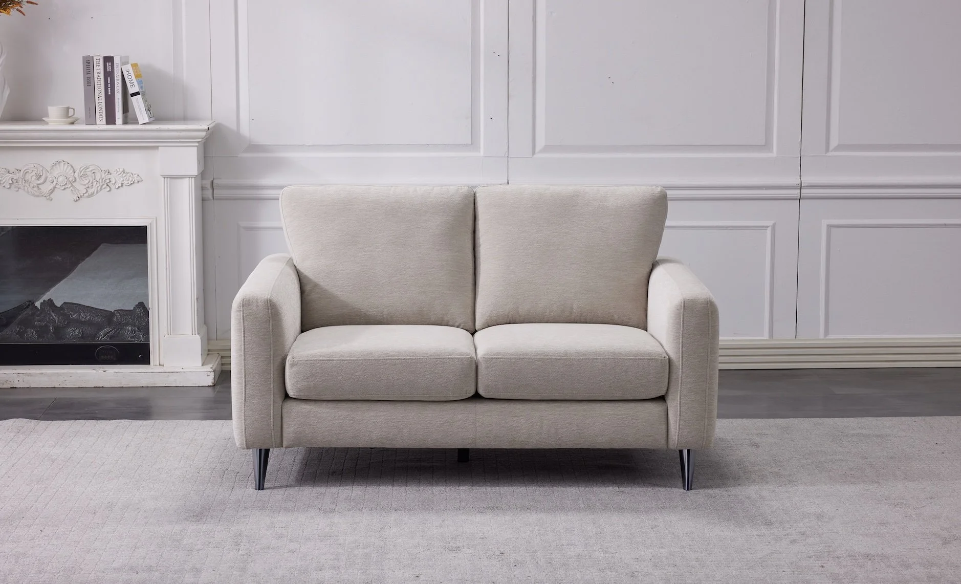 A light beige two-seater sofa with slightly angled backrests and thin black legs in a room with white paneled walls, a light-colored carpet, and a white ornate fireplace mantle with books and a small coffee cup on top.