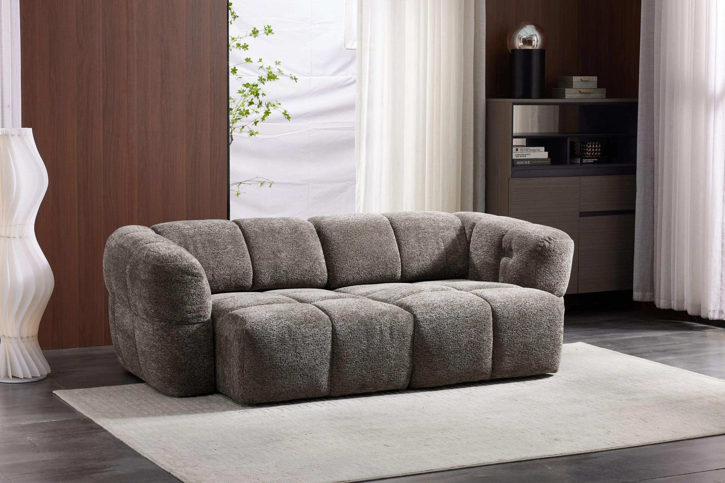 A modern living room with a gray upholstered sofa, a white textured lamp, a wooden wall, sheer white curtains, and a black and wood bookshelf with decorative items, all on a light-colored rug over dark flooring.