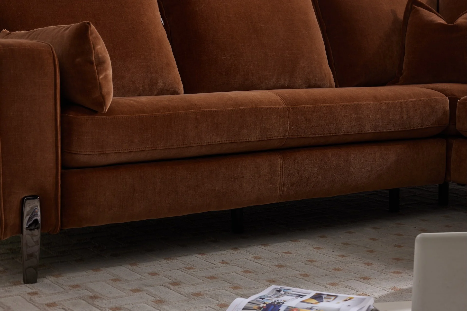 A brown fabric sofa with matching cushions on a beige patterned carpet, with a magazine and part of a white chair in the foreground.