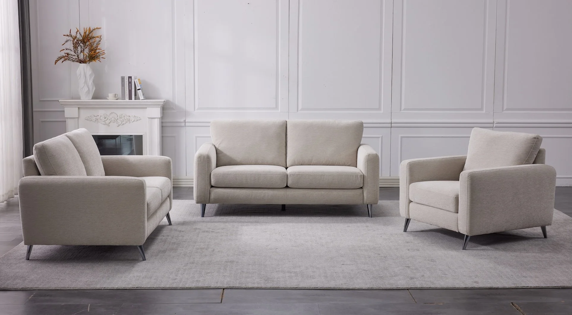 A minimalist living room with three beige upholstered sofas, a white decorative fireplace, a side table with books, and a vase with dried flowers on top. The room features white paneled walls and a light-colored area rug and dark hardwood floors.