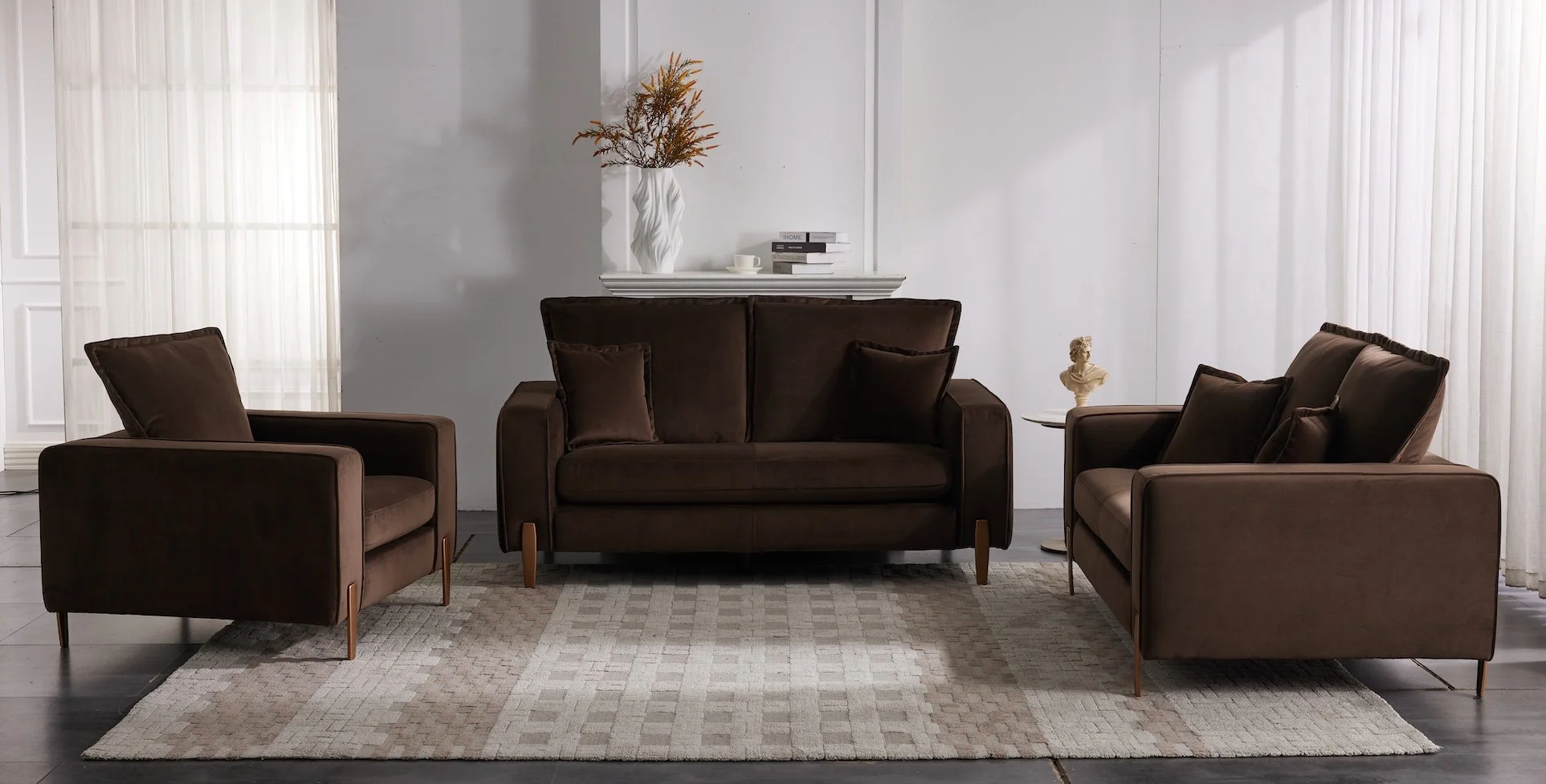 A living room with a dark brown sofa and two matching armchairs, a beige rug, a side table with a bust sculpture, and a shelf with books and a vase with dried flowers.