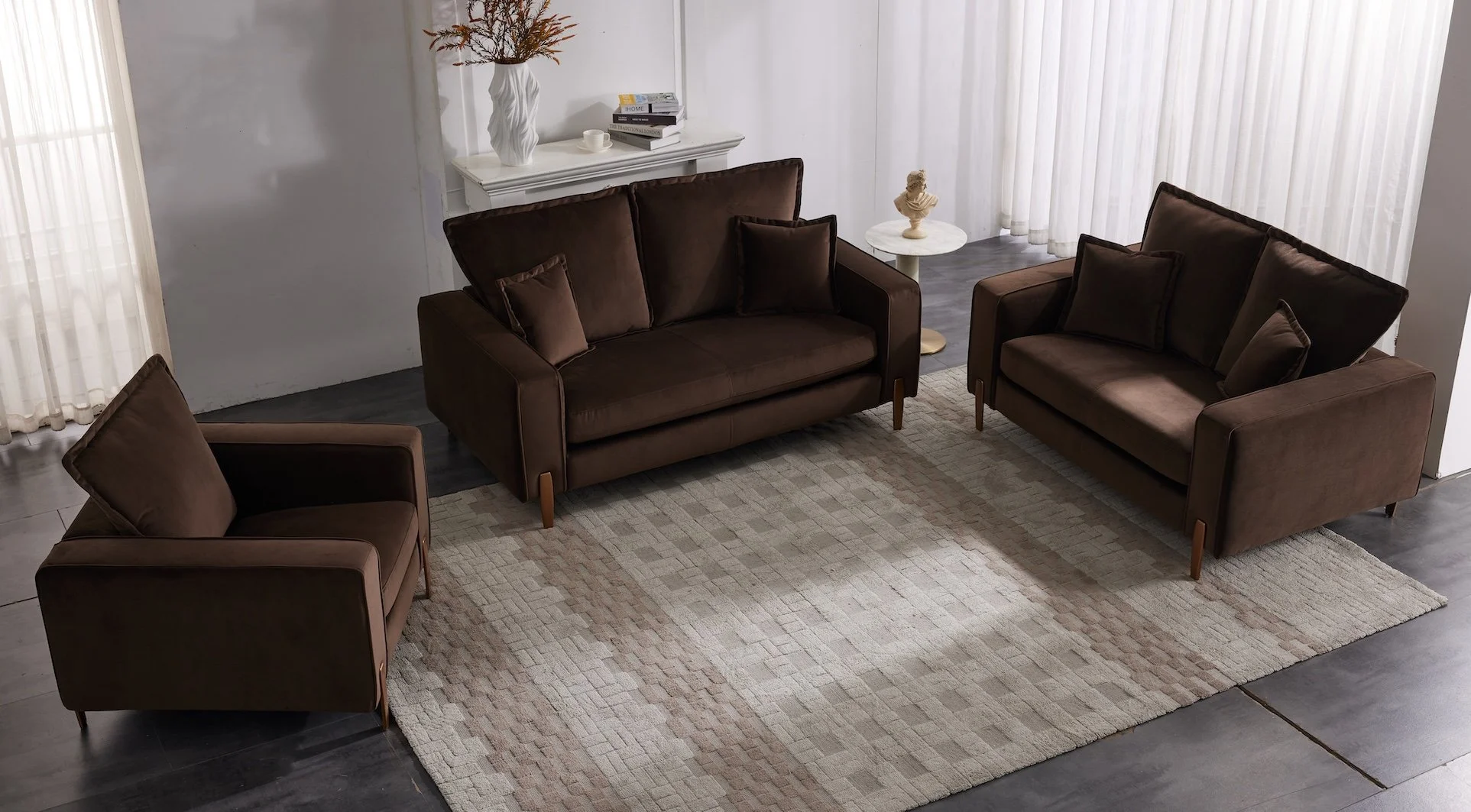 Three brown sofas with cushions arranged around a beige patterned rug in a living room with white curtains and a dark floor. There are decorative items and books on a table and a small side table with a bust sculpture.