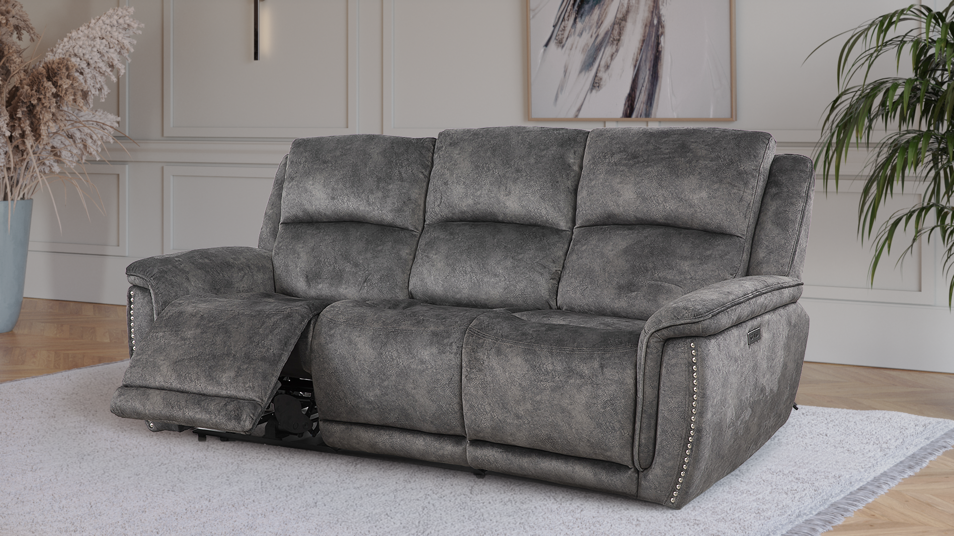 A modern living room with a white wall, large windows with sheer curtains, and dark hardwood floors. There is a dark gray three-seater sofa and two matching armchairs arranged around a beige area rug. A framed abstract art piece hangs on the wall above the sofa.