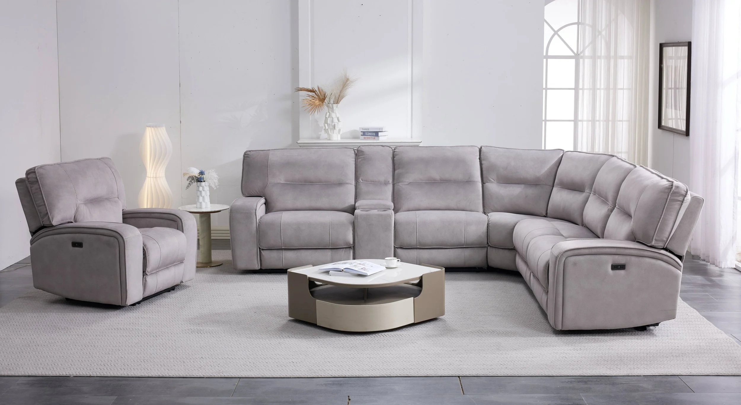 A modern living room with a beige sectional sofa, large arched windows with white curtains, a geometric mountain painting on the wall, a black side table with a small lamp and plant, and a patterned area rug on a dark wood floor.