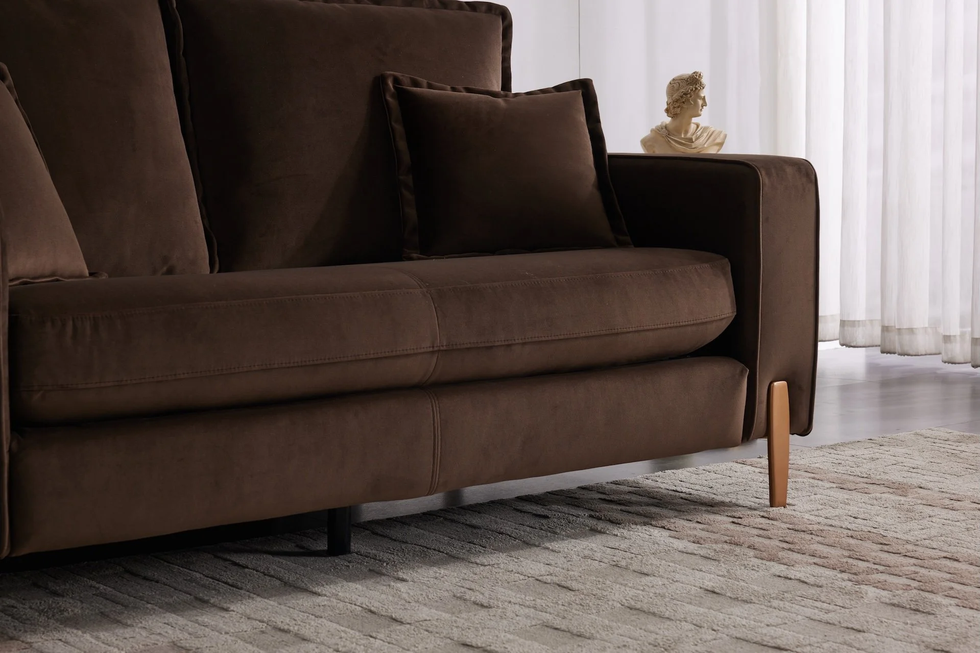 Close-up of a brown sofa with brown pillows, wooden legs, and a textured rug in a living room with white curtains and a bust sculpture in the background.