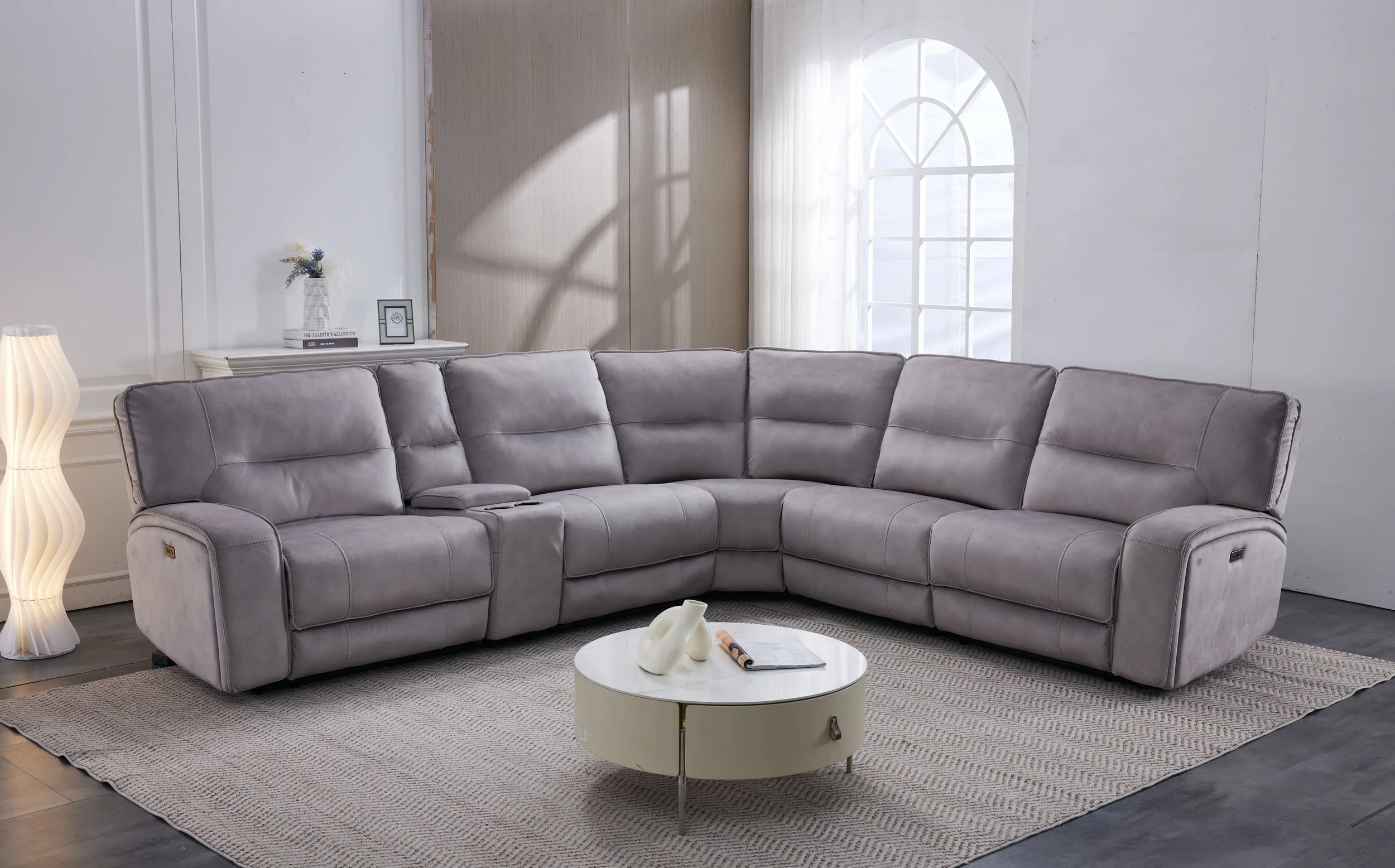 A modern living room with a beige sectional sofa, large arched windows with white curtains, a geometric mountain painting on the wall, a black side table with a small lamp and plant, and a patterned area rug on a dark wood floor.