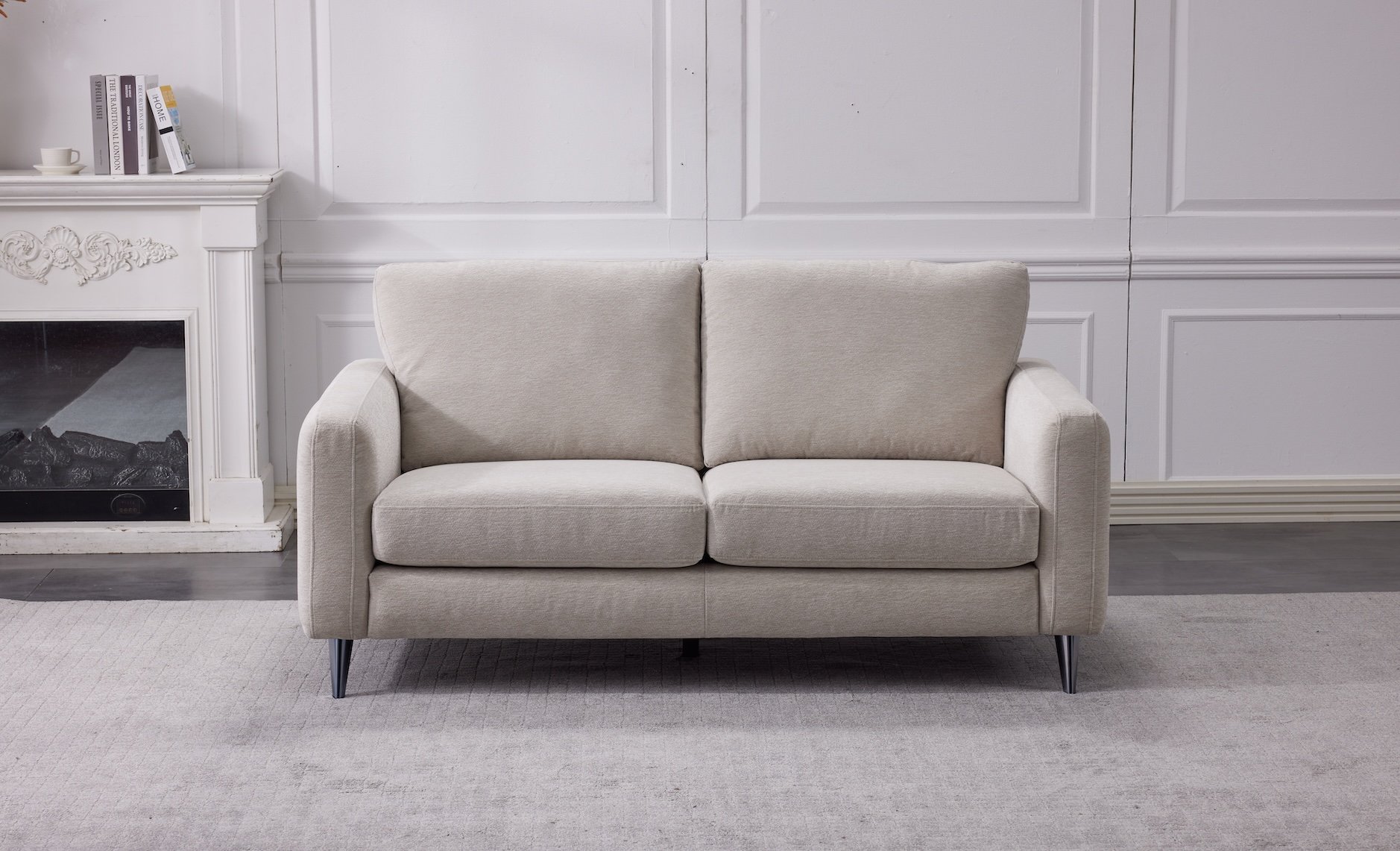 A beige two-seater sofa in a living room with white walls, a light gray carpet, and a white fireplace with books and a teacup on the mantel.