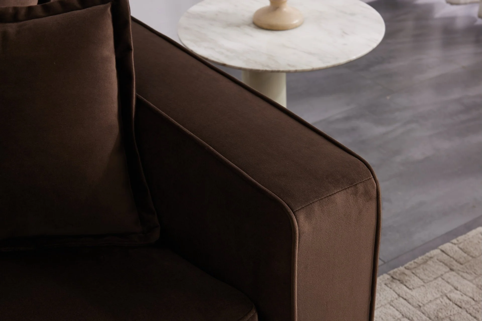 Close-up of a brown sofa with a matching cushion, next to a white marble side table with a beige lamp base, on a wooden floor with a rug.