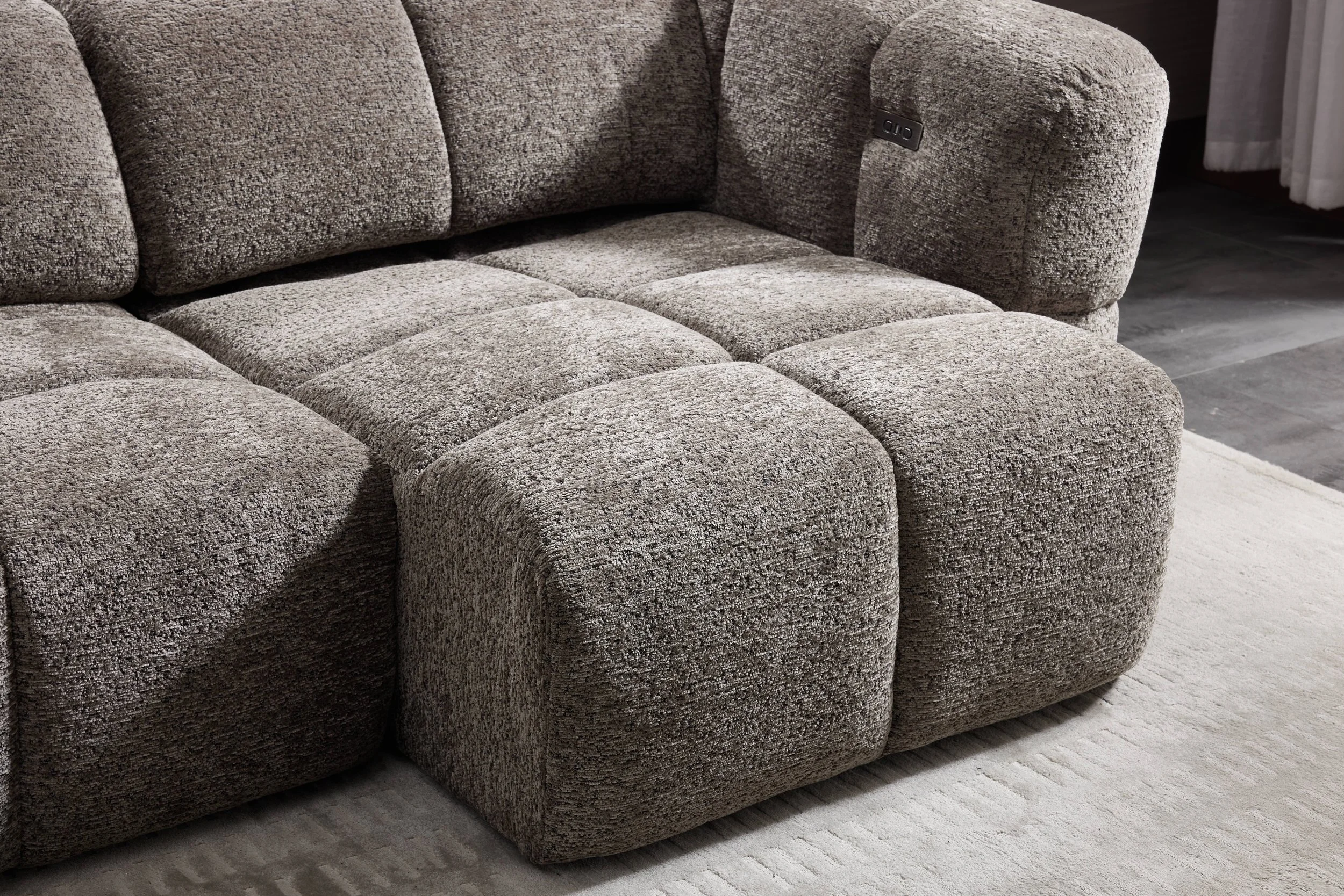 A close-up of a brown textured fabric sofa with plush cushions, situated on a light-colored rug in a room with dark flooring and curtains.