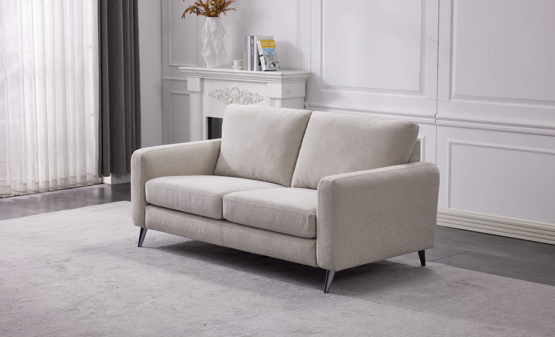 Light gray loveseat sofa in a modern living room with white walls, a white rug, and a fireplace mantle with books and a decorative vase.