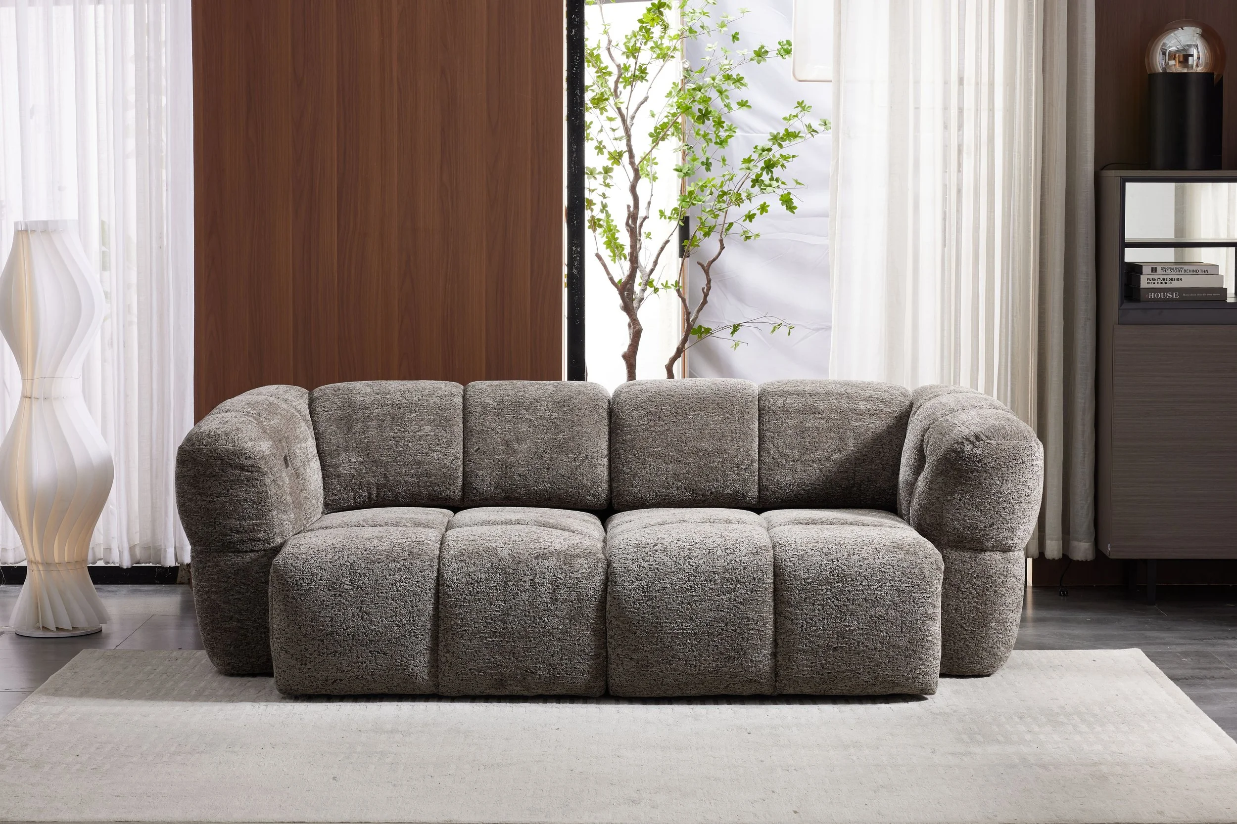 Living room with a modern gray upholstered sofa, a tall white sculpture lamp on the left, and a dark-colored cabinet on the right with books, a black decorative object, and a metallic sphere on top. Behind the sofa, there are sheer white curtains and