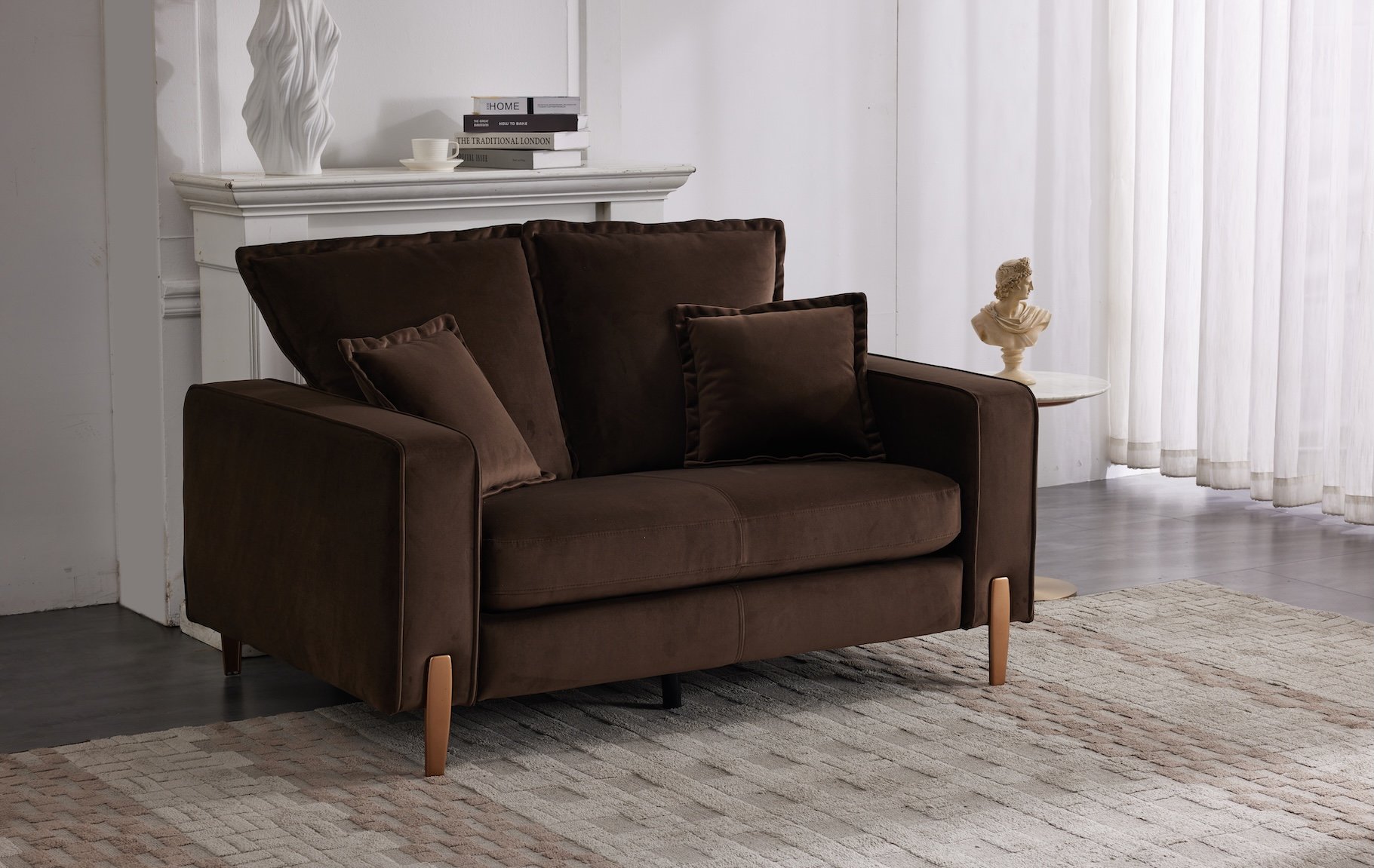 A living room corner with a dark brown sofa, matching cushions, a beige rug, a white fireplace mantel with a white sculpture, bowls, and books, a small round side table with a classical bust sculpture, and sheer white curtains letting in natural ligh