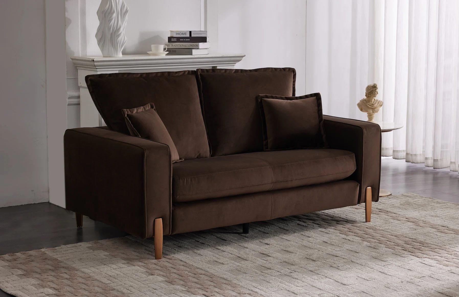 A brown sofa with matching cushions in a living room with a light rug, a white mantle with decorative objects and books on top, and white curtains covering a window.