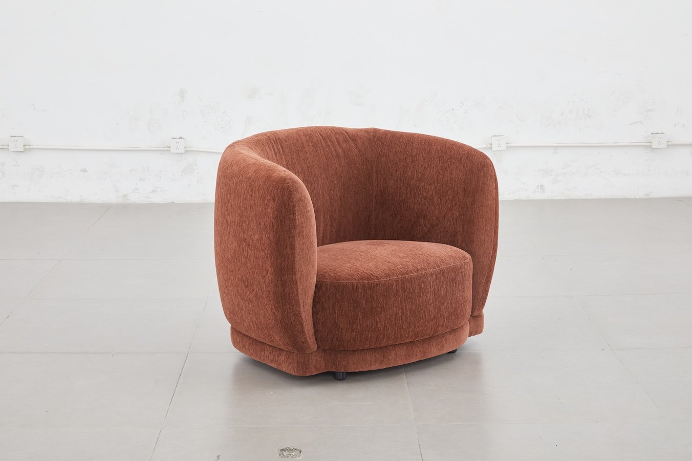 A rust-colored, upholstered, rounded armchair in a minimalist setting with white walls and light gray flooring.
