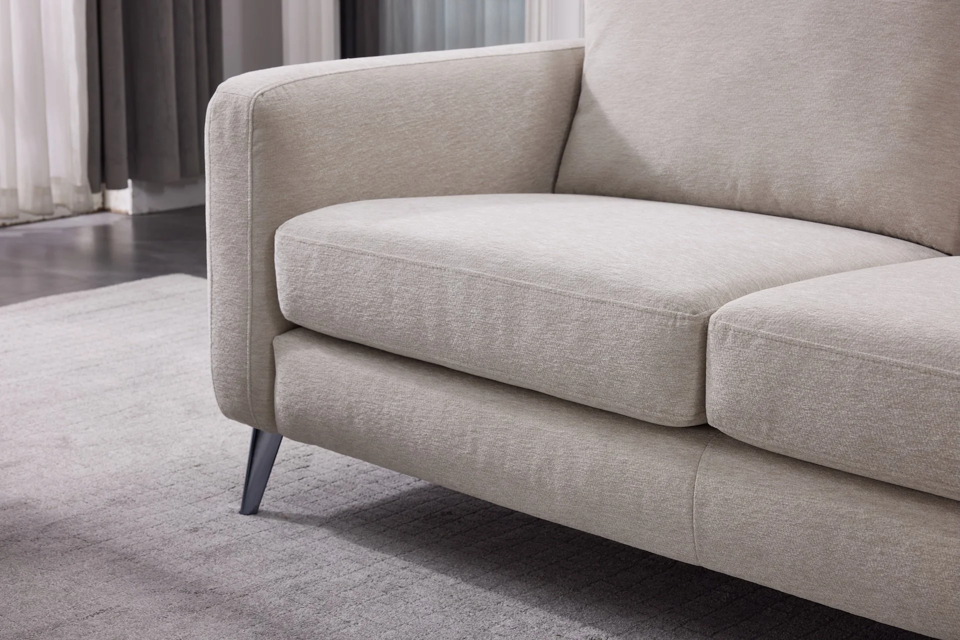 Close-up of a beige modern sofa with angled dark legs on a light textured area rug in a living room with gray curtains in the background.