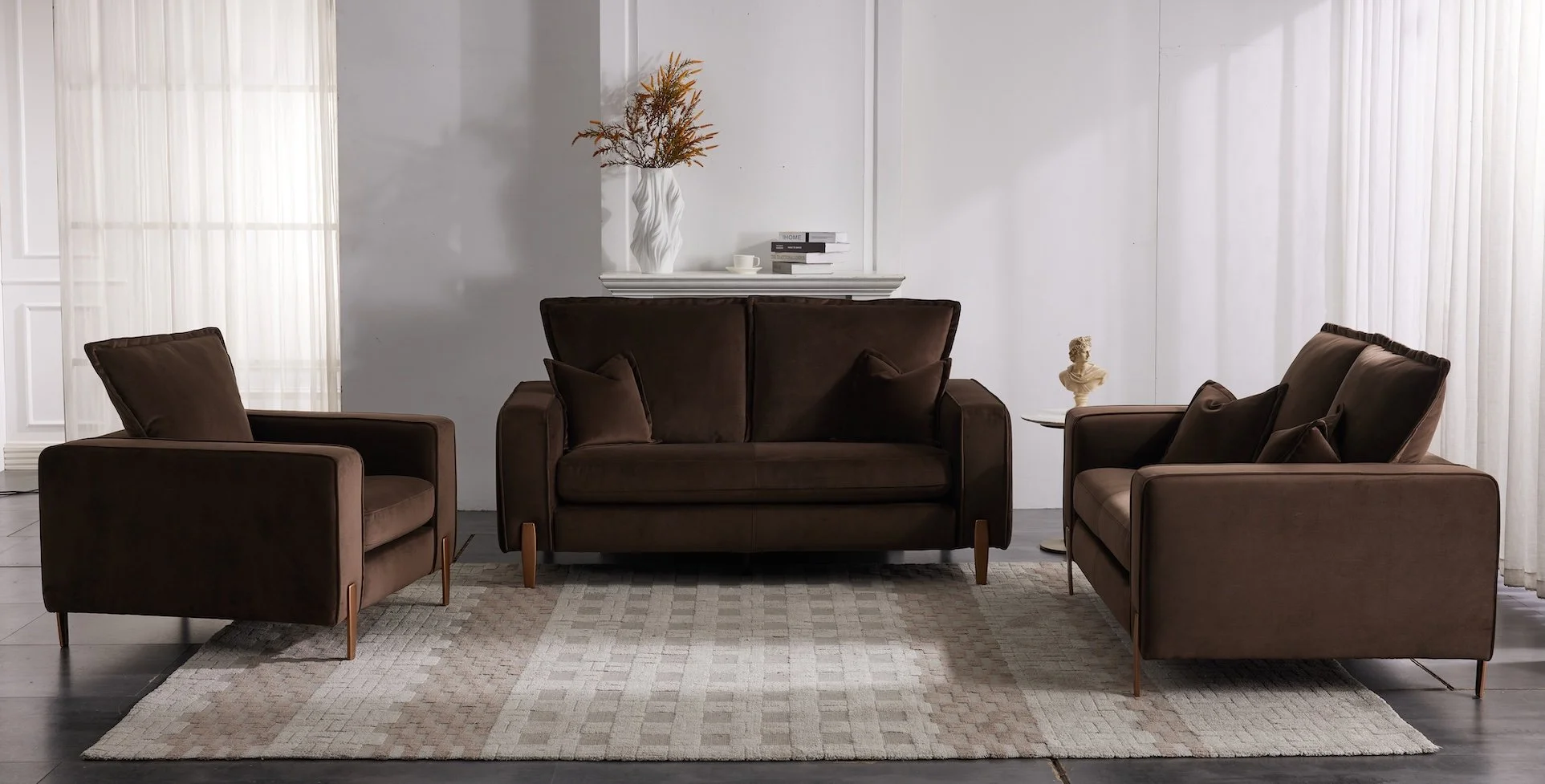 Living room with a brown sofa, two matching armchairs, a beige rug, white curtains, and decorative items including a vase with branches, a small bust, and stacked books on a shelf.