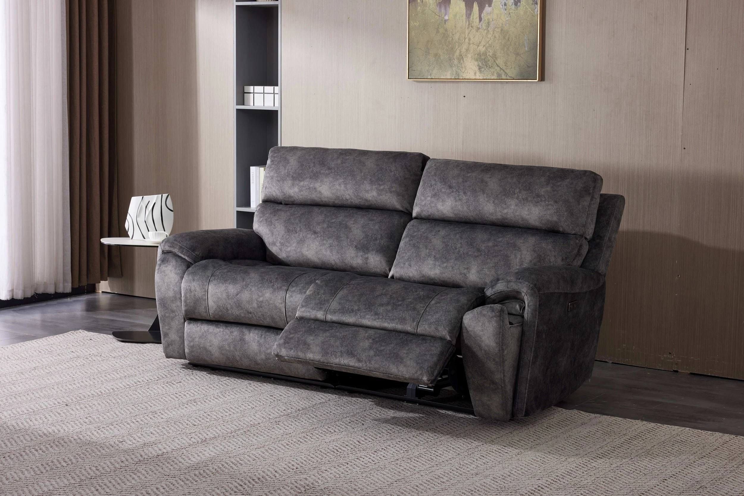 A modern living room with a white wall, large windows with sheer curtains, and dark hardwood floors. There is a dark gray three-seater sofa and two matching armchairs arranged around a beige area rug. A framed abstract art piece hangs on the wall above the sofa.