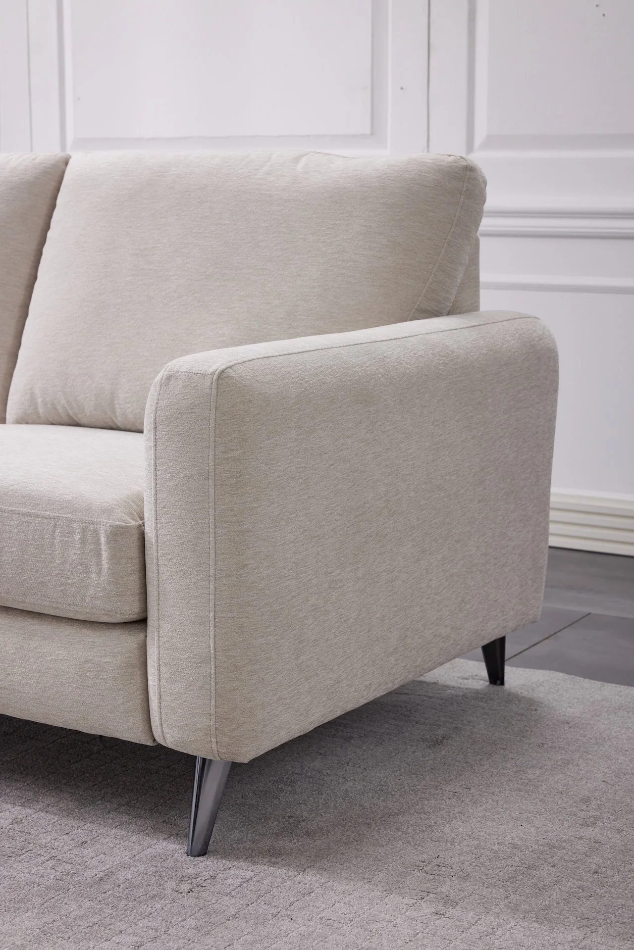 Close-up of a beige sofa with black tapered legs, placed on a light gray carpet in a room with white walls and wainscoting.