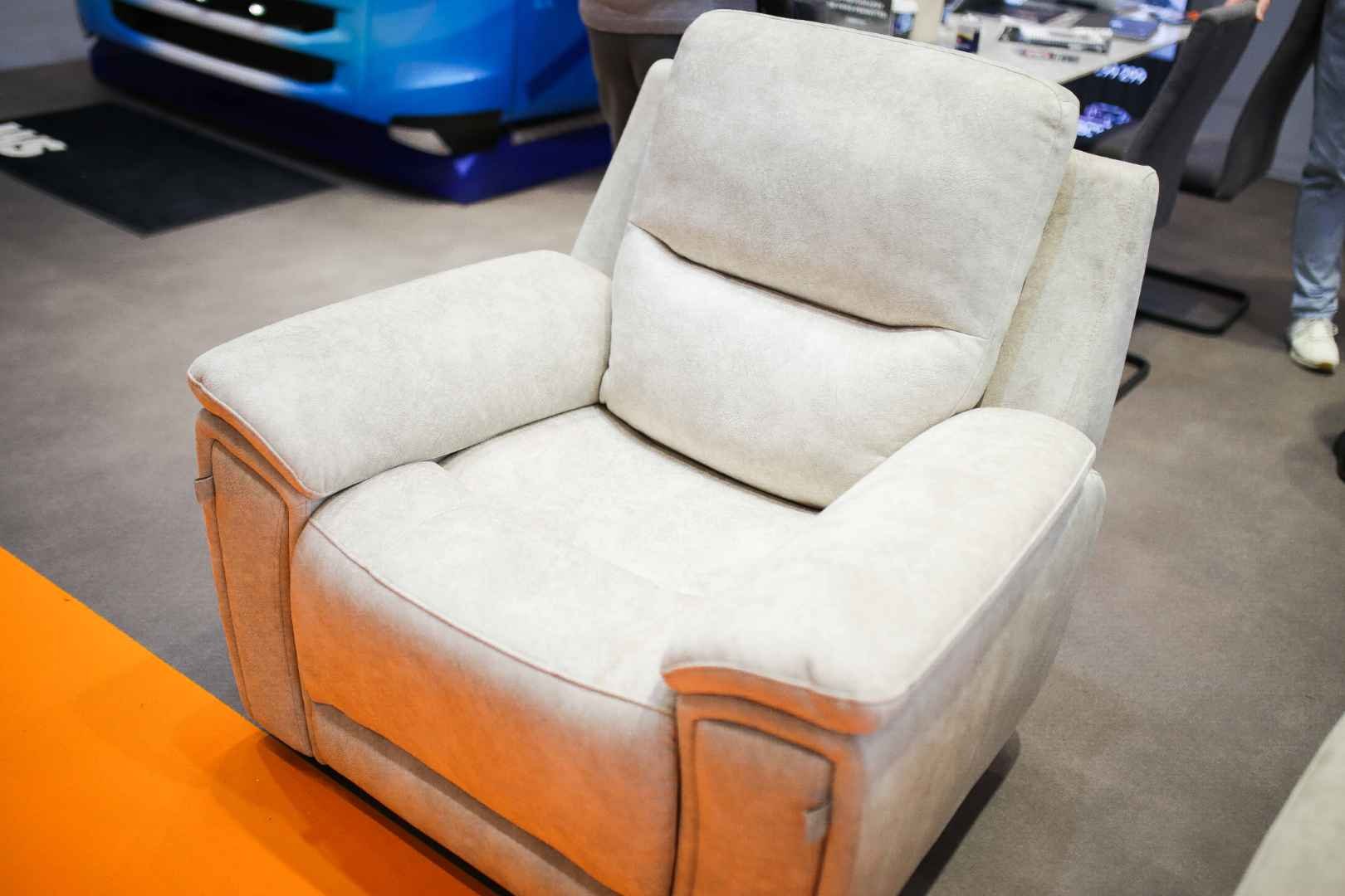 A beige upholstered armchair with padded armrests and a matching back cushion, displayed on a showroom floor.